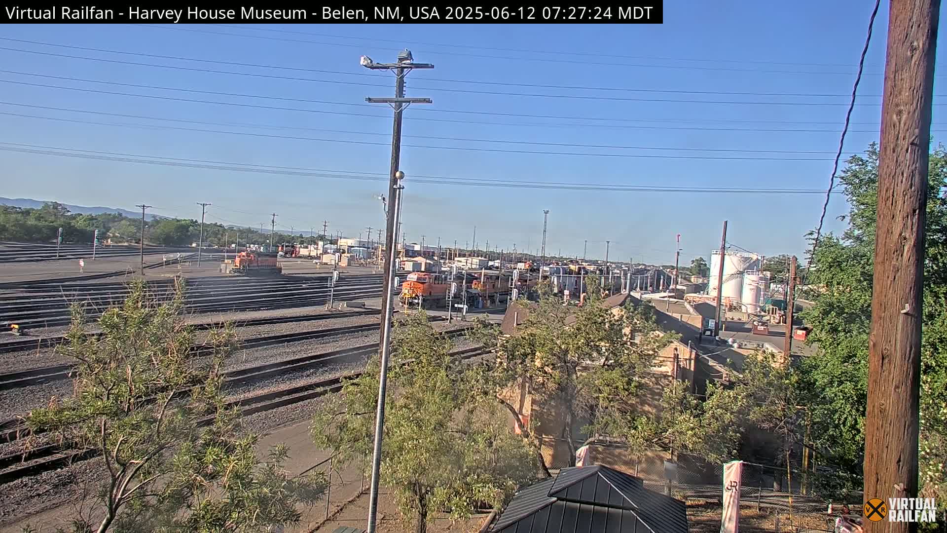 A sunny day at a rail yard shows multiple train cars and locomotives, some orange, situated along numerous train tracks with surrounding vegetation and buildings visible in the distance.