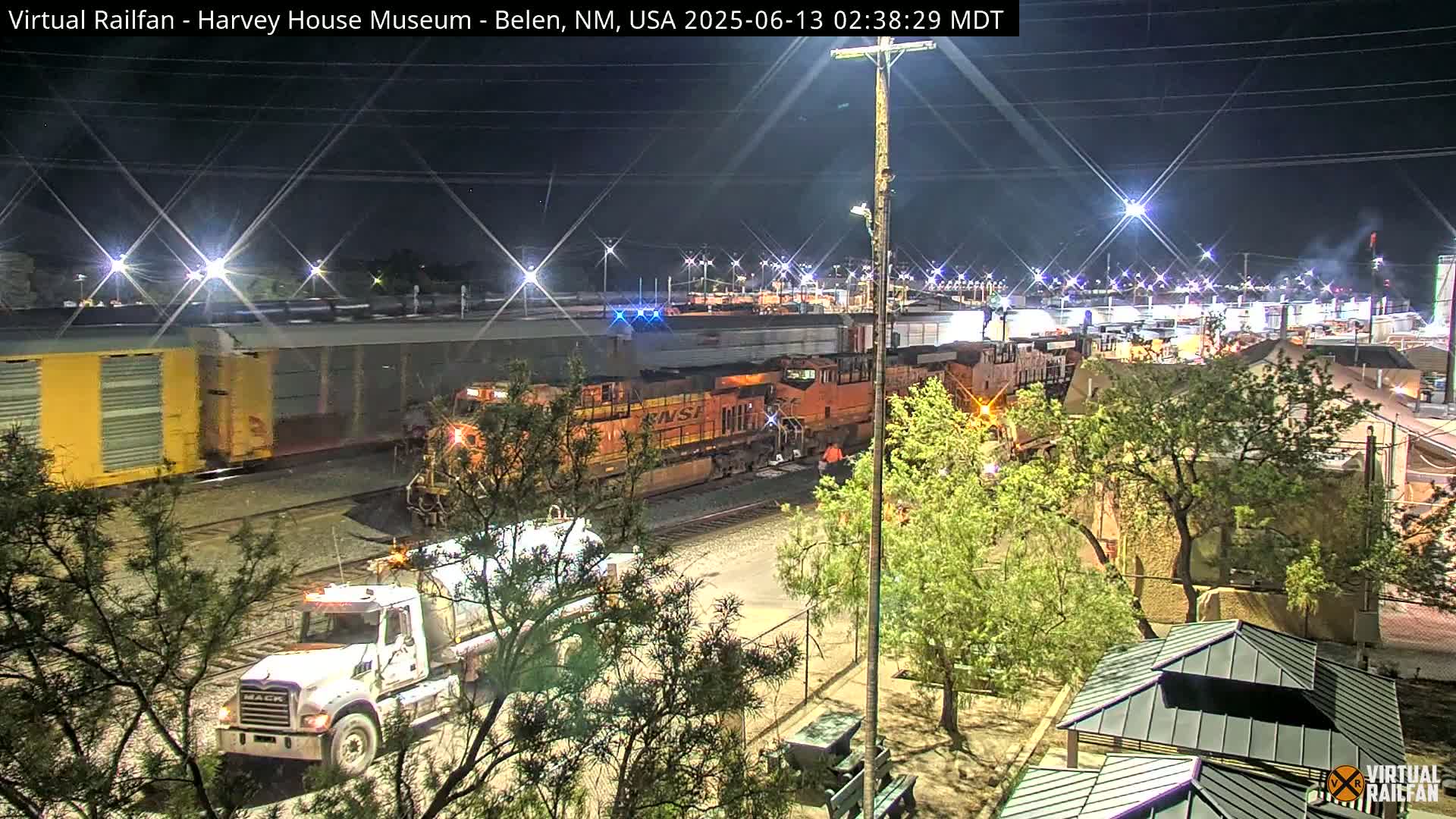 At night, a train with two orange locomotives is parked next to a white tanker truck in a rail yard with surrounding trees and lights.