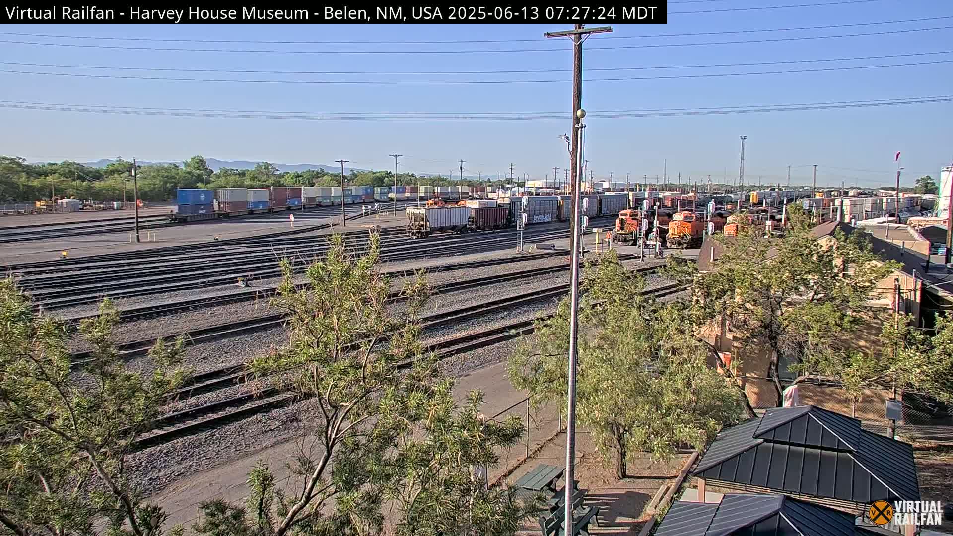A sunny day at a rail yard with numerous train cars and locomotives, surrounded by trees and a building.