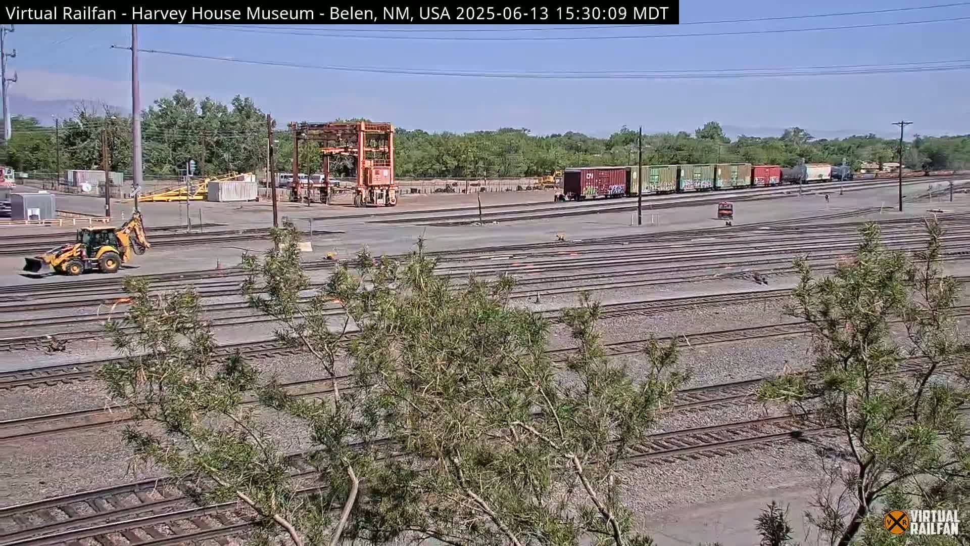 A yellow excavator sits near a train yard with many parallel tracks under a clear, sunny sky.
