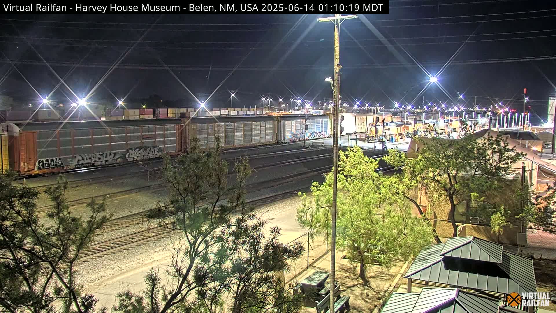 A nighttime view of a railway yard with several boxcars, some displaying graffiti, under bright artificial lighting, surrounded by trees and a small park-like area.