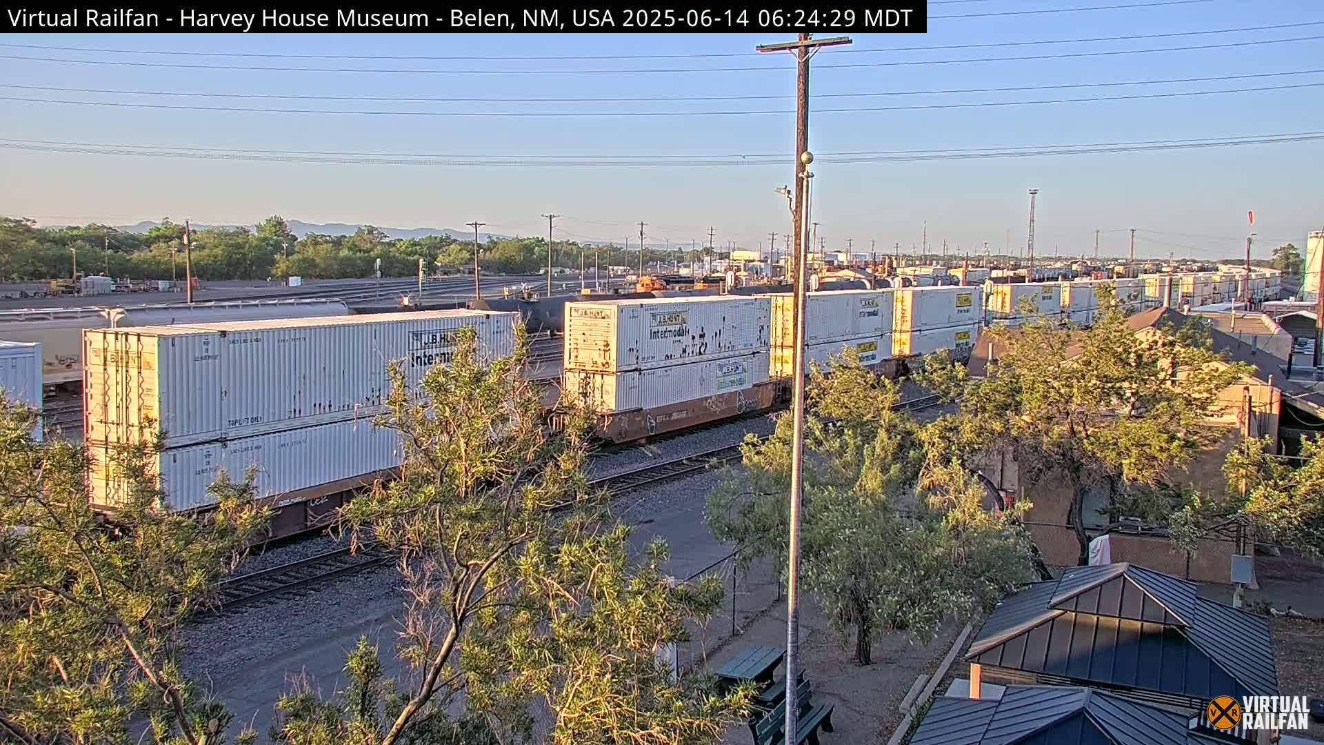 A long freight train carrying numerous shipping containers is visible under a clear, bright sky, with some trees and buildings in the foreground.