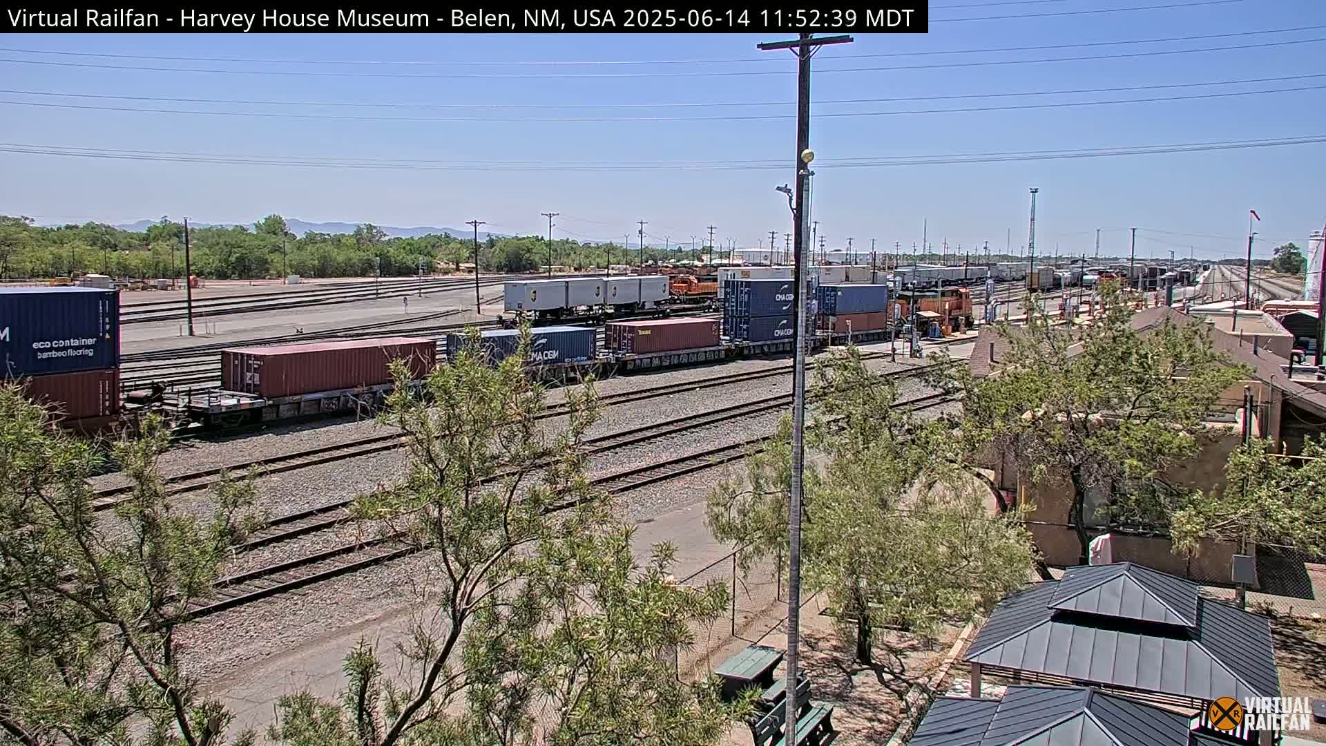 Under a clear, sunny sky, a train yard with numerous cargo containers sits on multiple parallel train tracks, partially obscured by trees and a building.