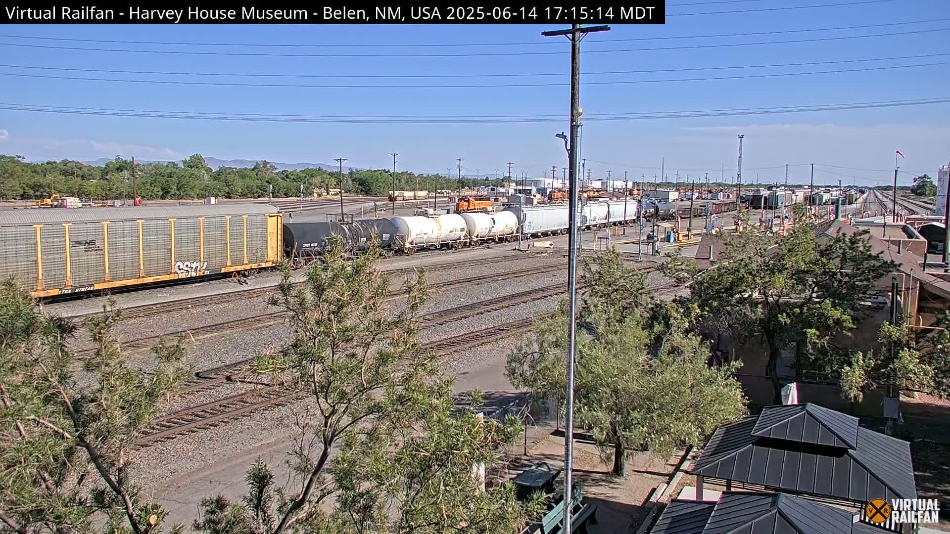 Under a clear, sunny sky, a train yard with numerous cargo containers sits on multiple parallel train tracks, partially obscured by trees and a building.