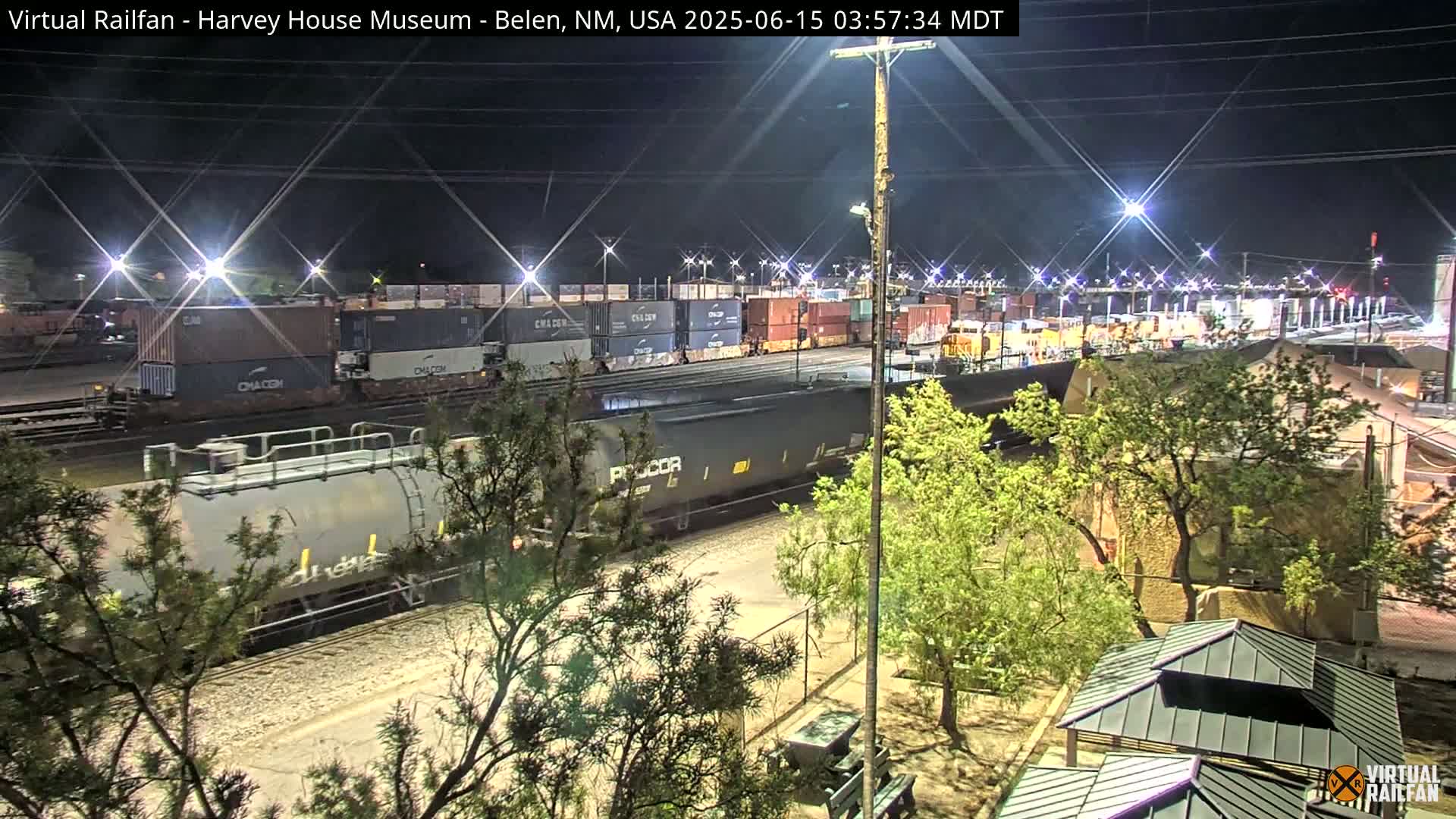 At night, a freight train passes by a rail yard filled with shipping containers, next to a park with trees and covered picnic structures under bright lights.