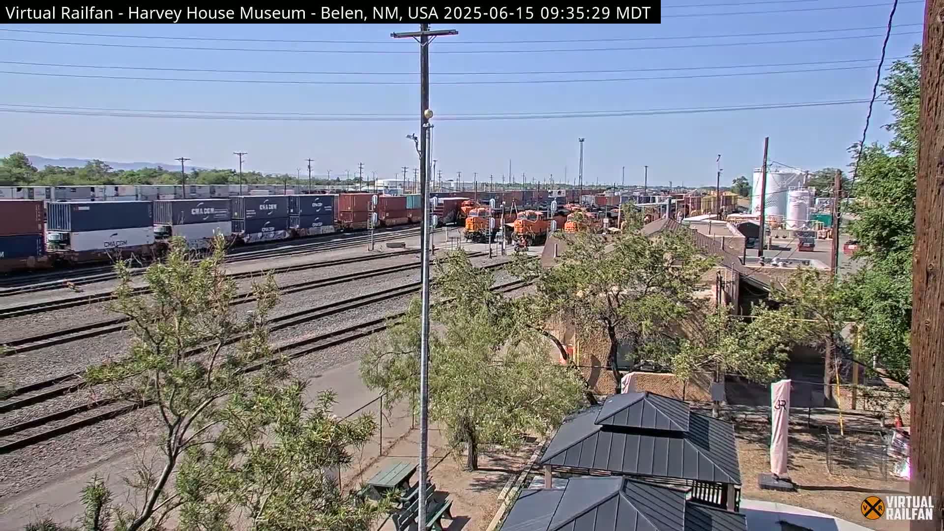 A sunny day at a railway yard shows several orange locomotives and freight cars with shipping containers, alongside trees, buildings, and park-like structures.