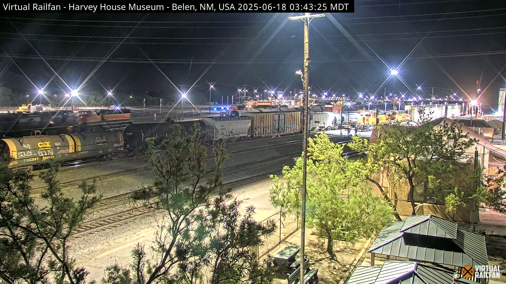 A nighttime view of a train yard with several freight cars, illuminated by lights, surrounded by trees and a small building.
