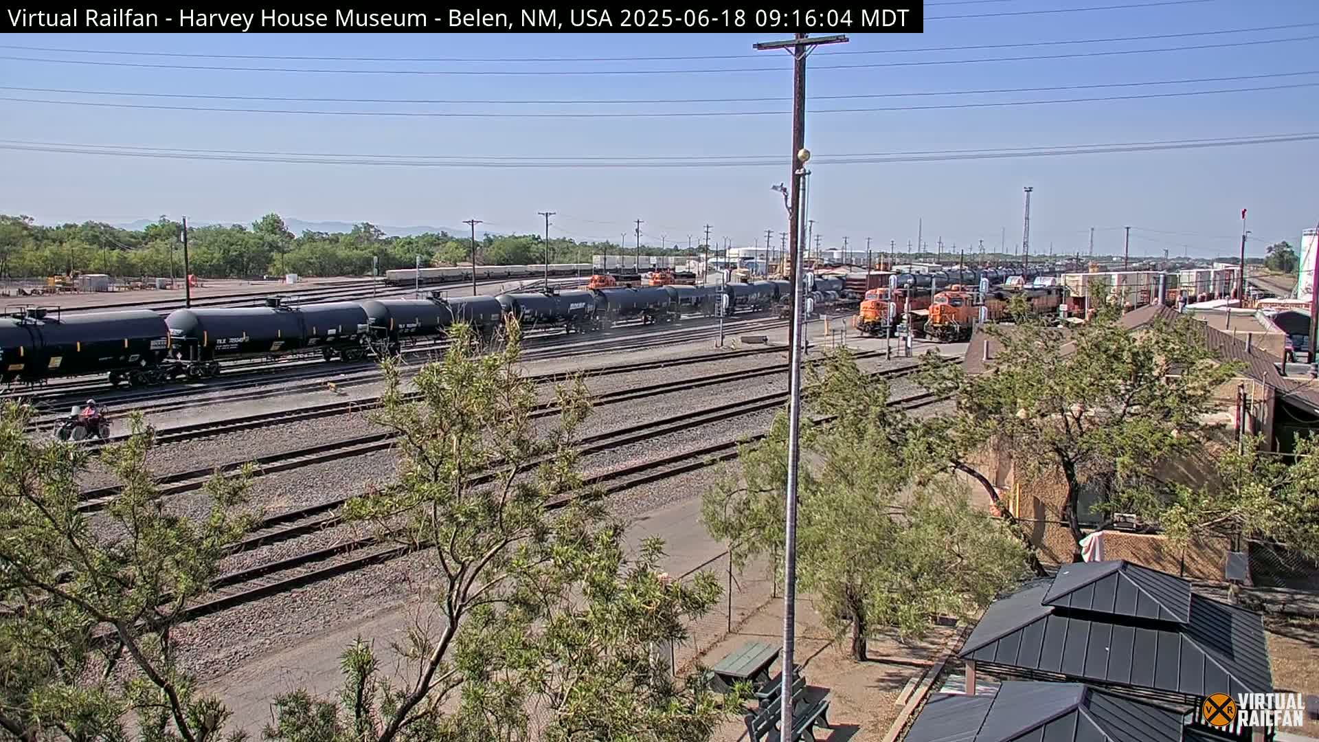 A sunny day reveals a railroad yard with several tanker cars and locomotives, surrounded by trees and a few buildings.