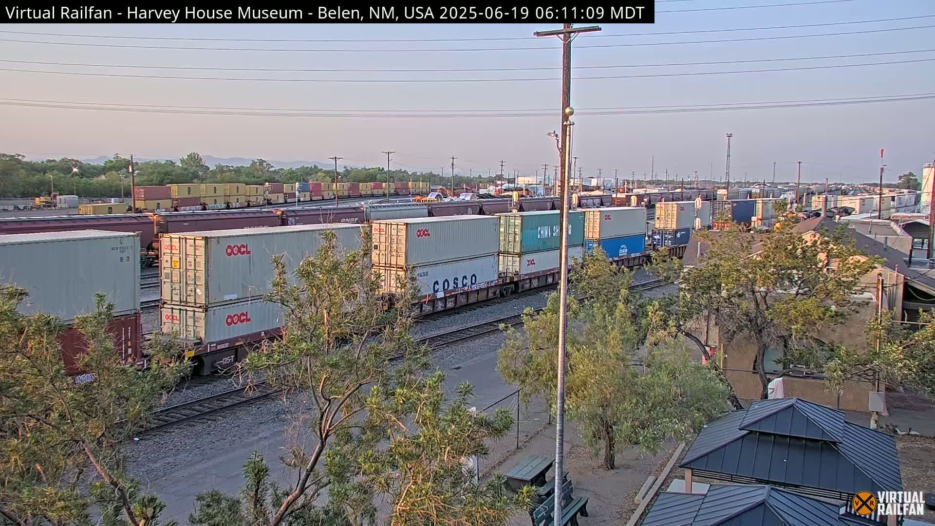 A long freight train carrying multiple shipping containers is visible on railway tracks under a clear, light sky.