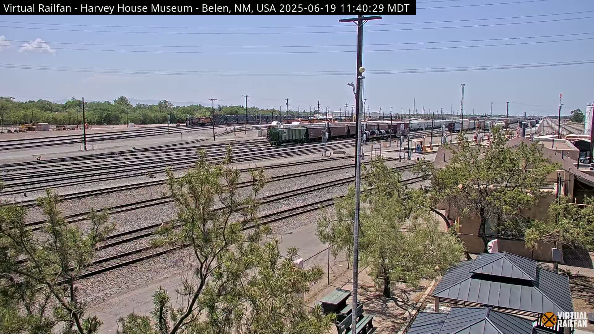 A sunny day reveals a wide expanse of railroad tracks and yards filled with freight cars, alongside sparse vegetation and a low building.