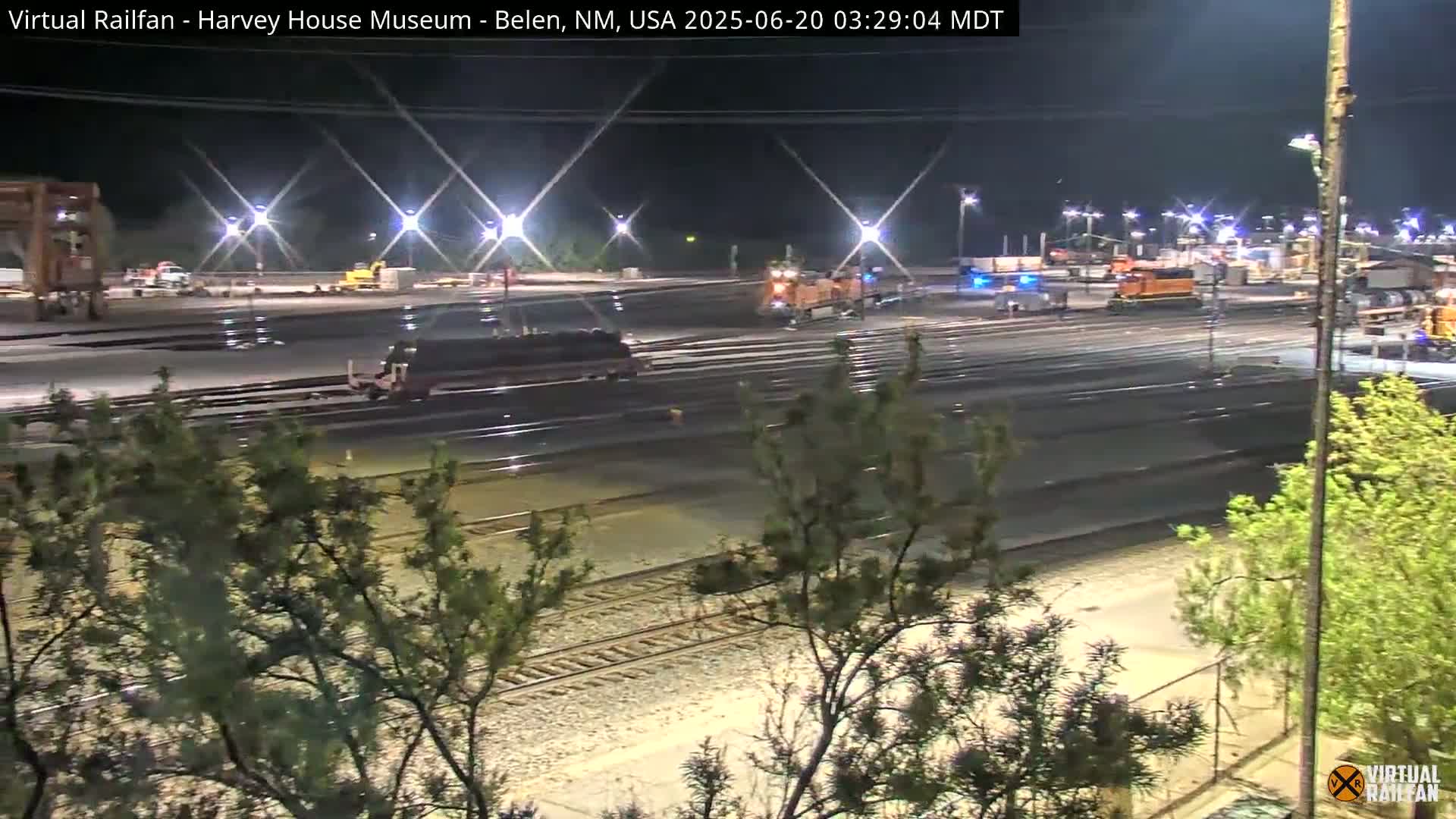 A nighttime view of a rail yard with several pieces of rail equipment and some vegetation in the foreground.
