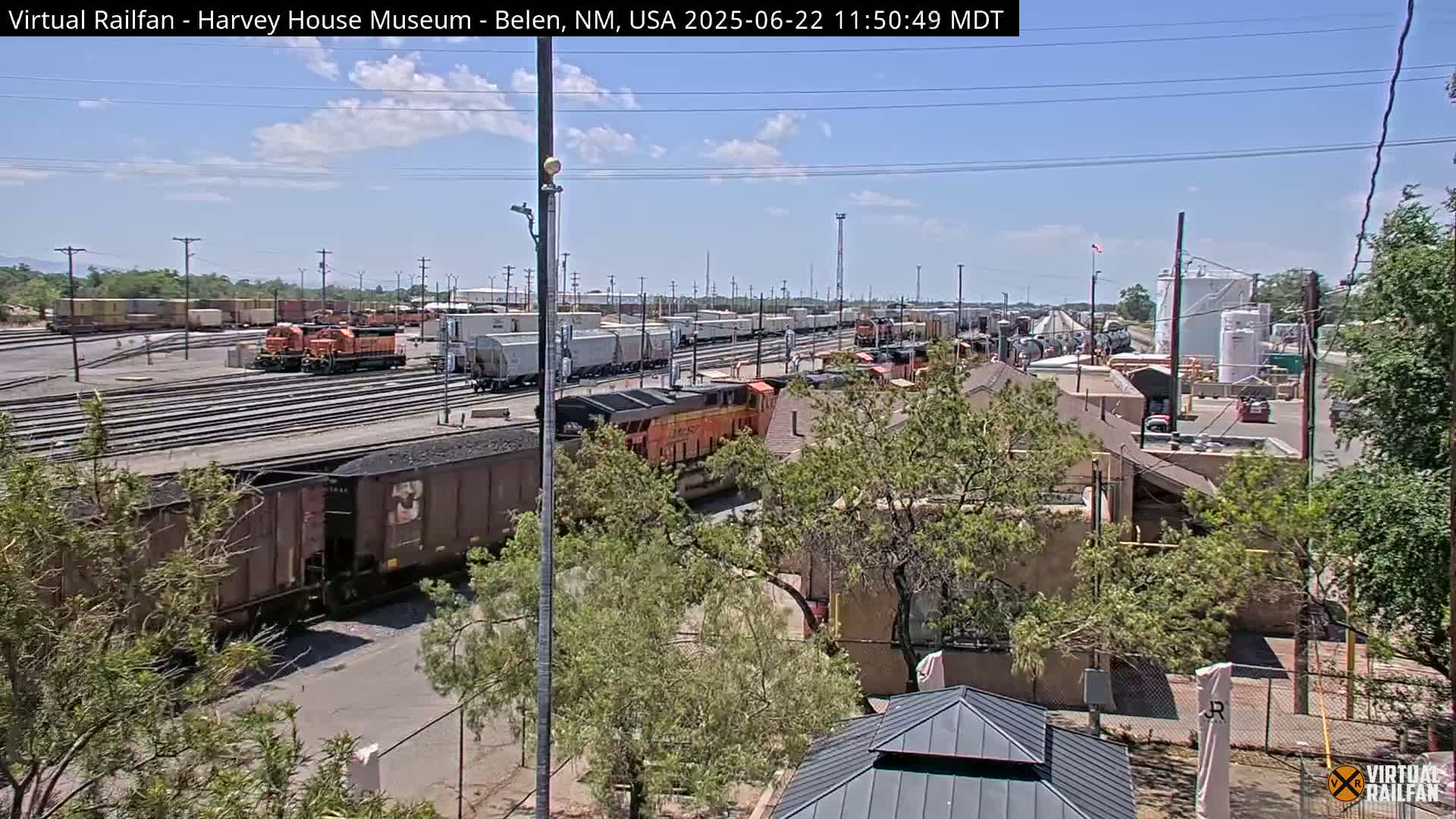A sunny day reveals a railroad yard with numerous train cars and locomotives, some orange, others brown, along with industrial buildings and lush green vegetation.