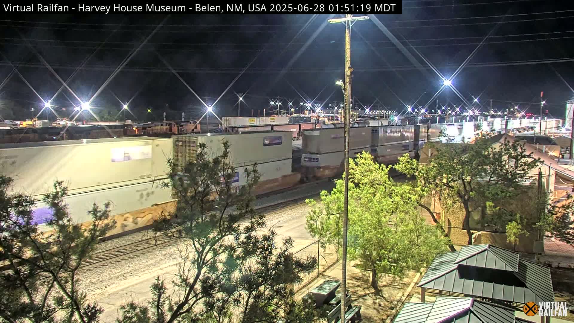 A nighttime view of a freight train with multiple shipping containers passing through a rail yard, under bright artificial lighting.