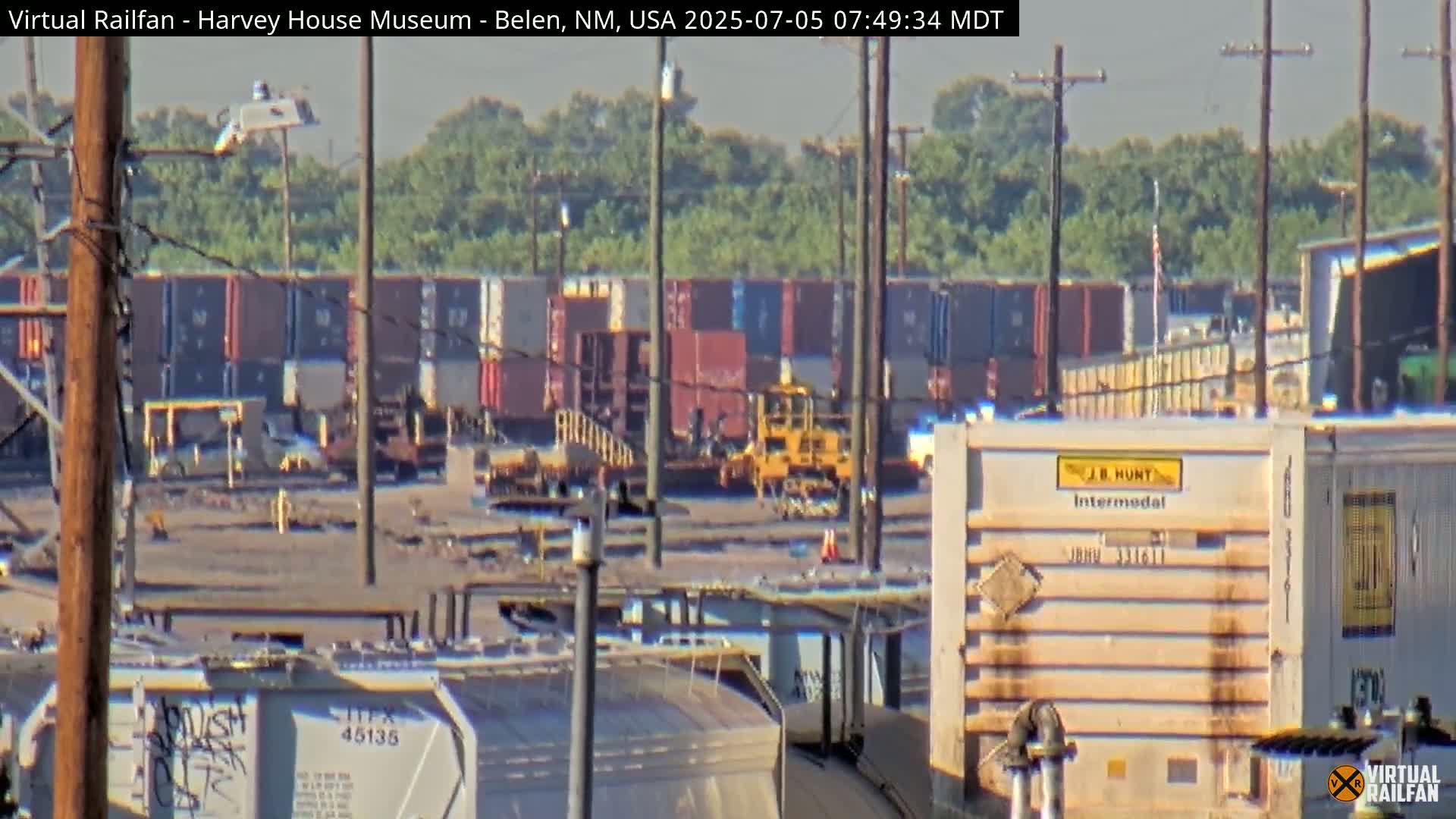 A hazy, sunny day reveals a train yard filled with numerous freight containers and railcars, with a yellow piece of railway equipment visible in the mid-ground.