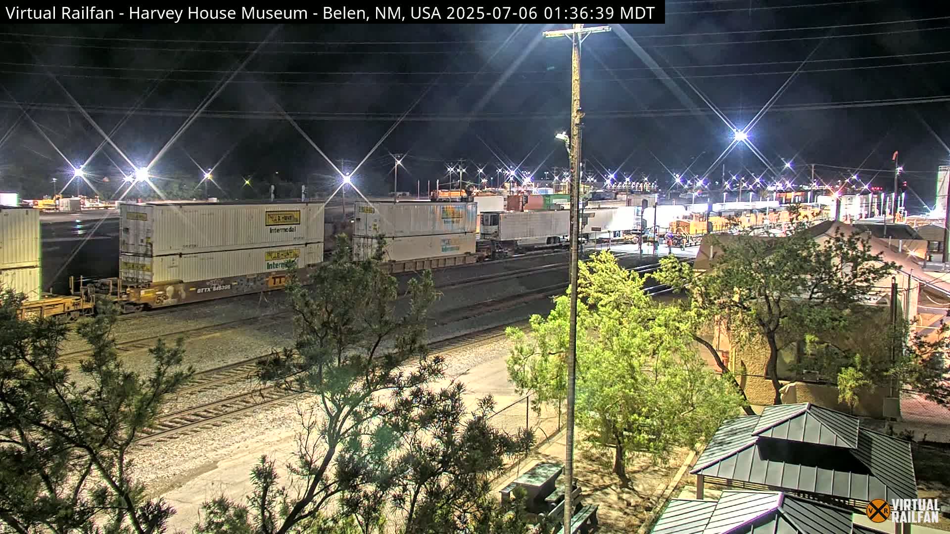 A nighttime view of a train carrying shipping containers in a rail yard, illuminated by artificial lights, with trees and buildings visible in the surrounding area.