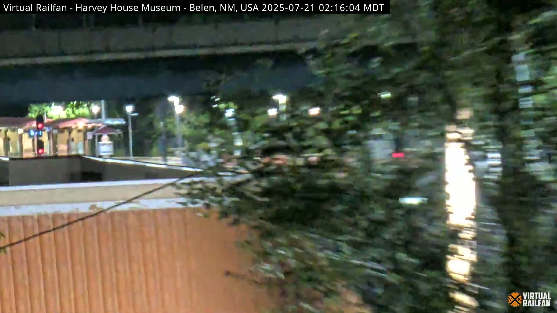 A nighttime view of a train station partially obscured by dense foliage, with lights illuminating the scene.