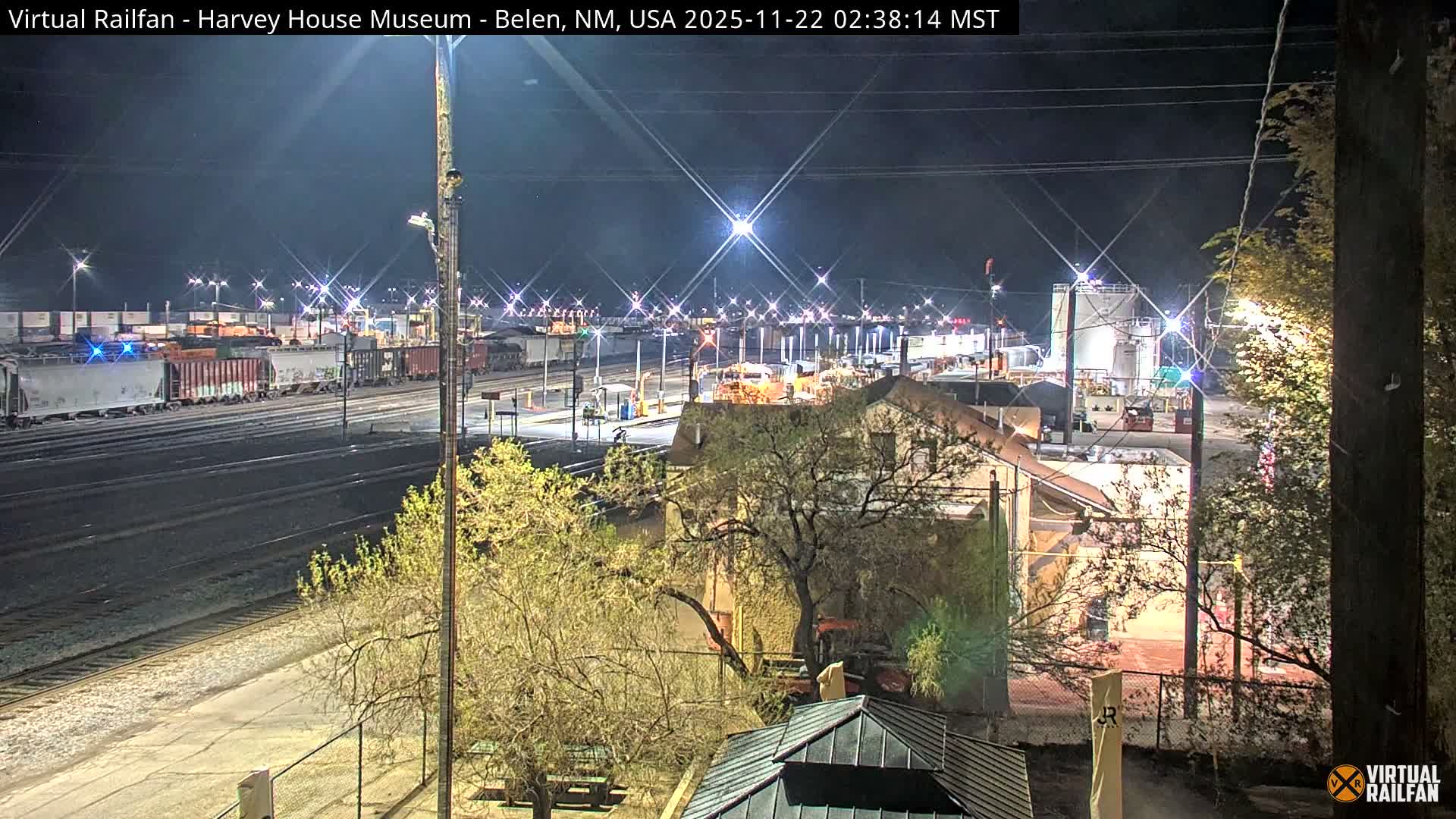 A nighttime aerial view captures a busy rail yard with multiple tracks and a long freight train, flanked by buildings, industrial structures, and bare trees under a clear, dark sky.