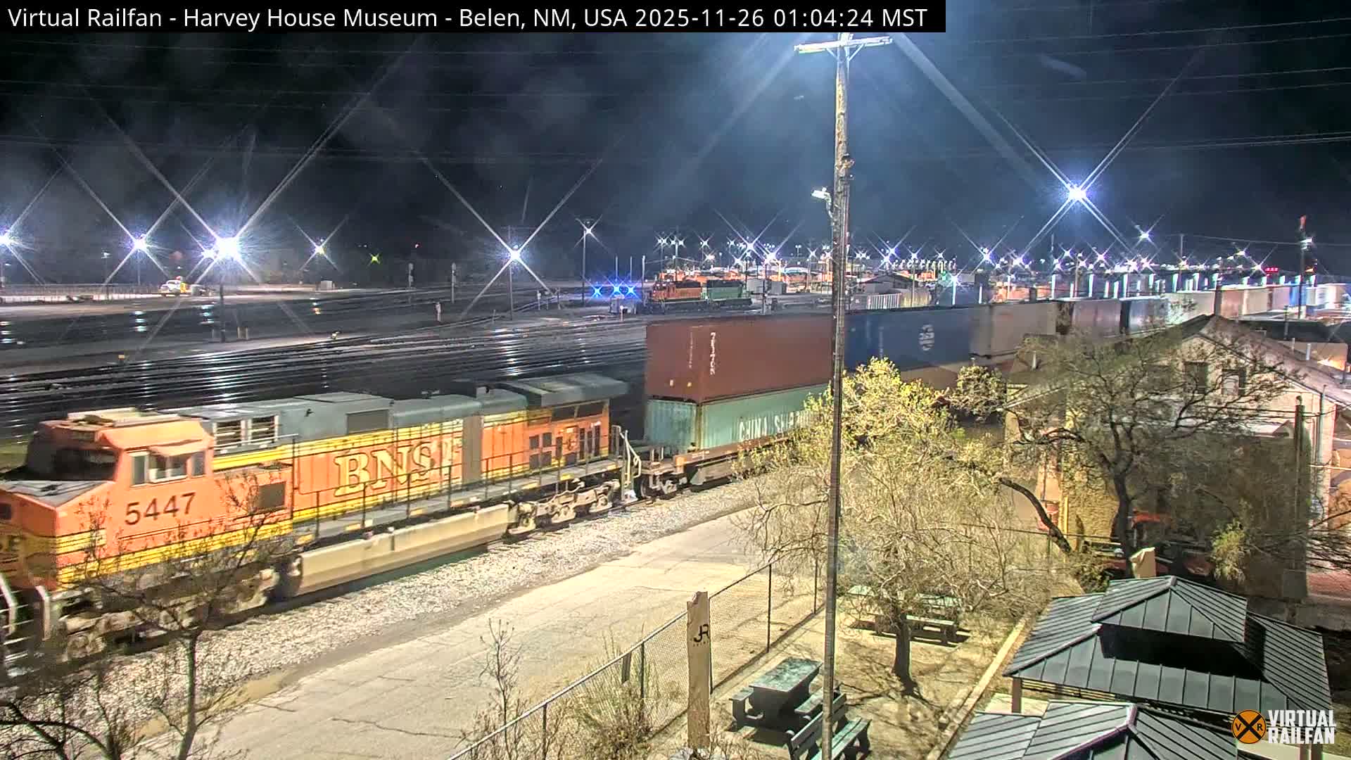 A long orange and yellow freight train with numerous container cars moves through a brightly illuminated rail yard at night under clear weather conditions, with multiple tracks, bare trees, and buildings visible in the scene.