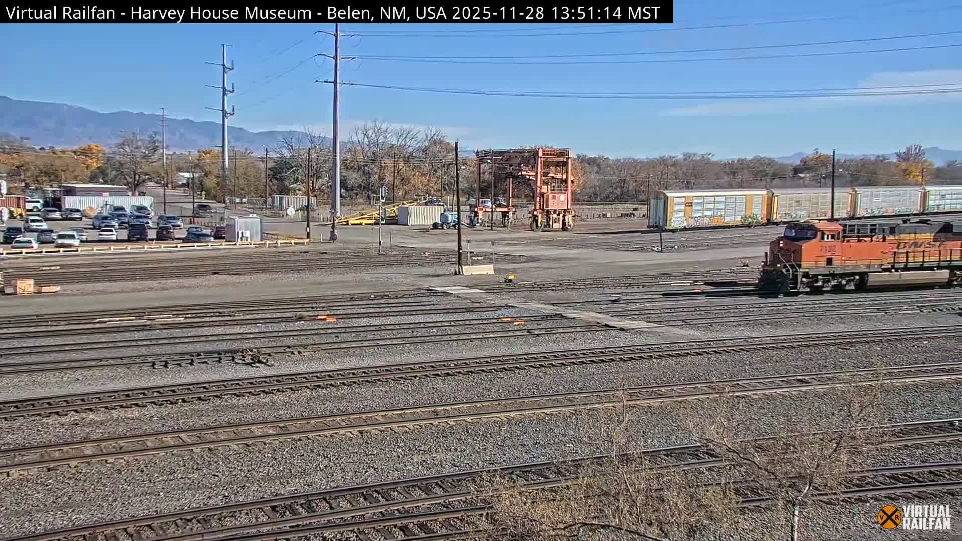 An orange BNSF train is visible in a large rail yard with multiple tracks, industrial equipment, and parked cars, all under a clear blue sky on a sunny day with distant mountains in the background.