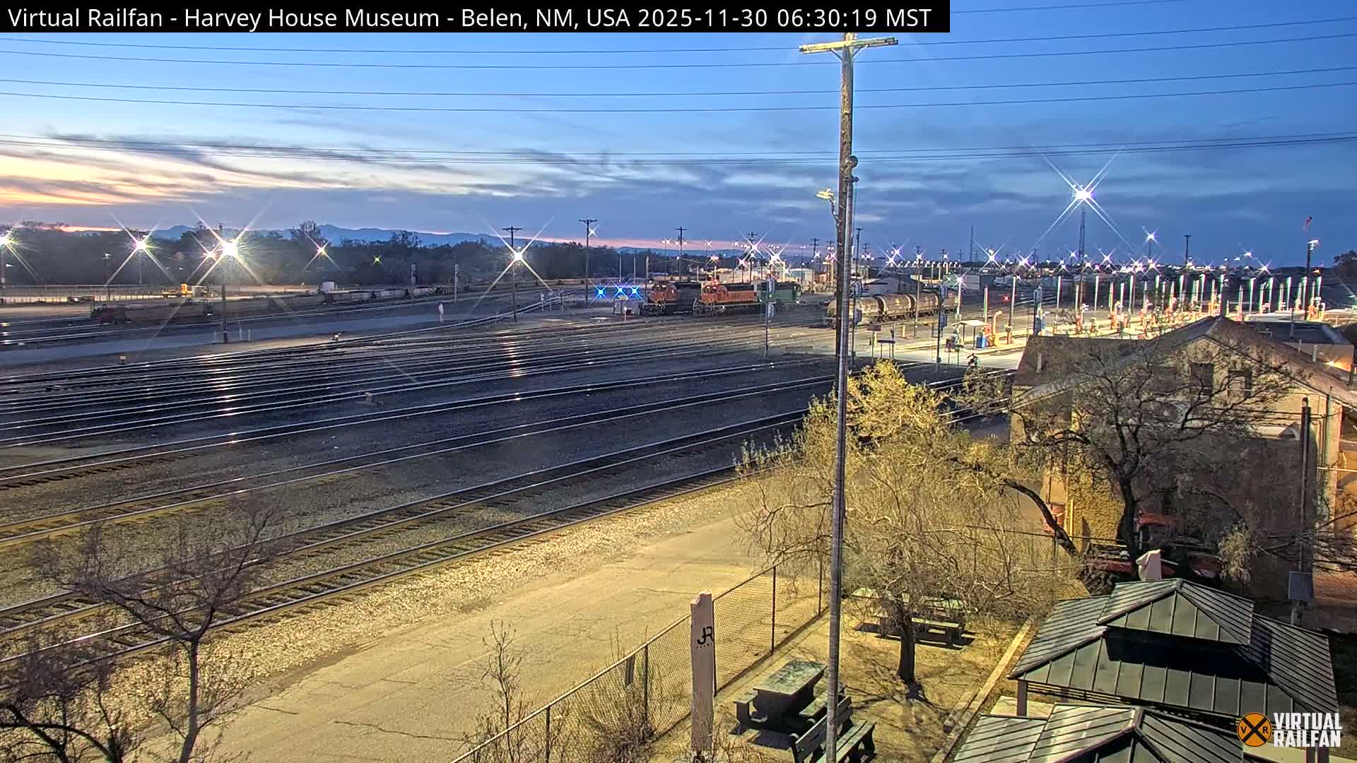 An orange BNSF train is visible in a large rail yard with multiple tracks, industrial equipment, and parked cars, all under a clear blue sky on a sunny day with distant mountains in the background.