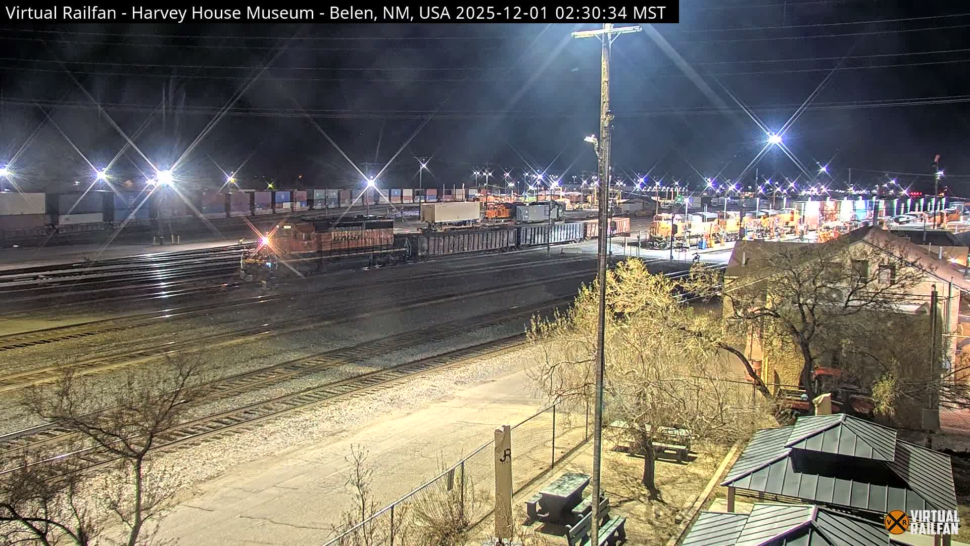 Under a clear, dark night sky, a wide train yard featuring numerous tracks and freight trains is brightly illuminated by starburst streetlights, with a building and leafless trees occupying the right foreground.