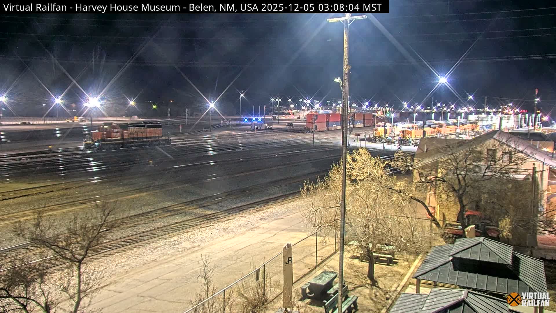 From an elevated perspective, a brightly lit train yard with multiple tracks, a locomotive, shipping containers, and industrial facilities is visible under a clear night sky, with a building and bare trees in the foreground.