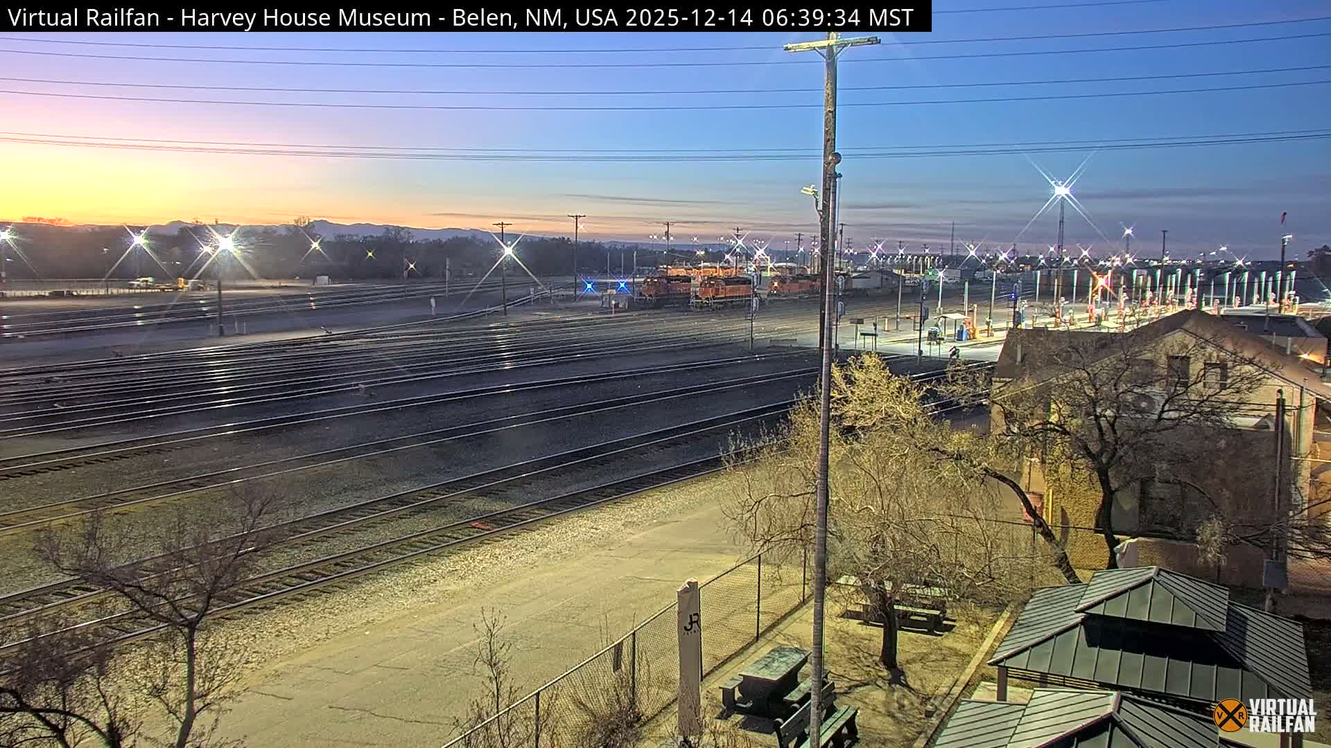 From an elevated perspective, a brightly lit train yard with multiple tracks, a locomotive, shipping containers, and industrial facilities is visible under a clear night sky, with a building and bare trees in the foreground.