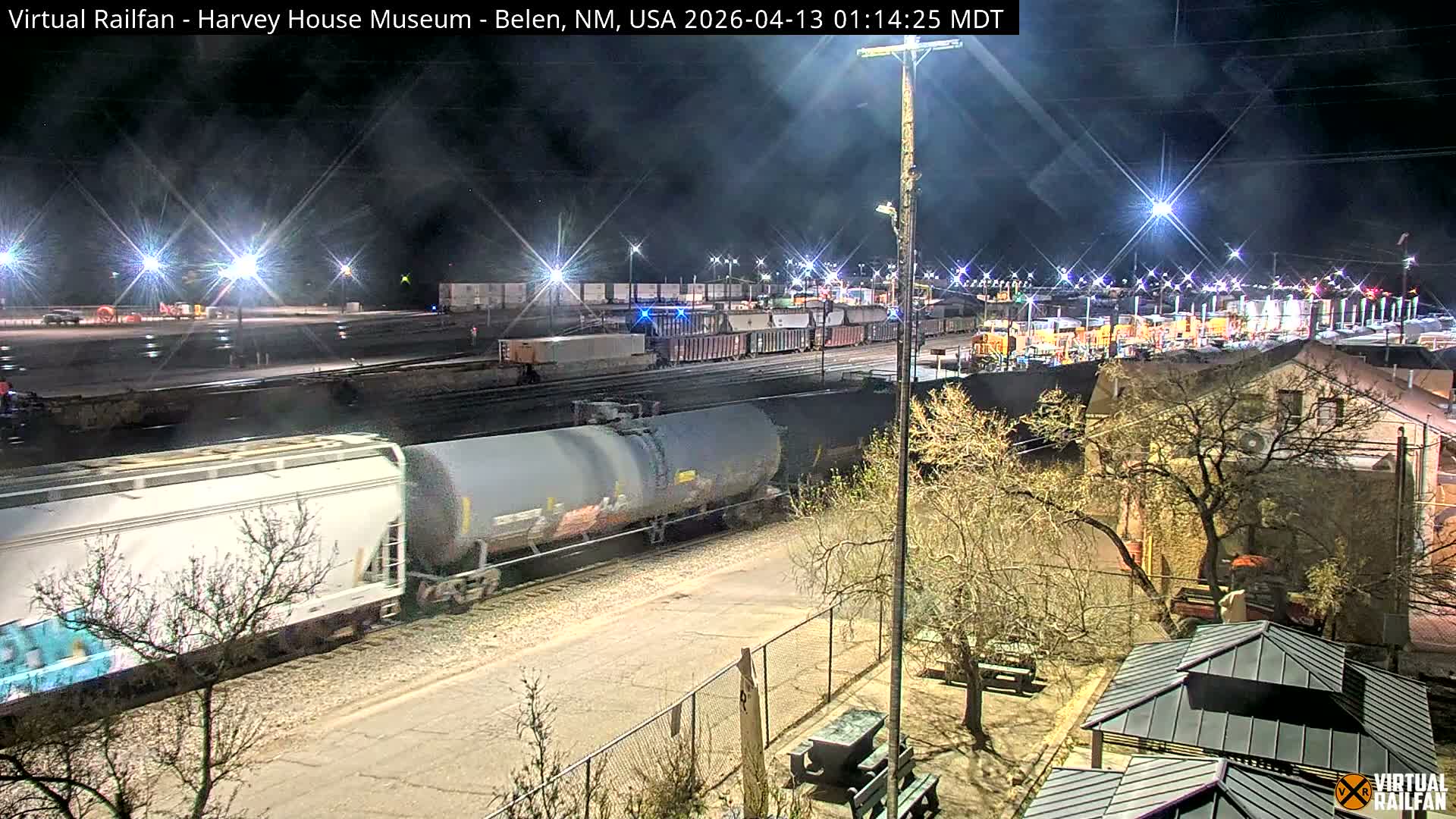From an elevated perspective, a brightly lit train yard with multiple tracks, a locomotive, shipping containers, and industrial facilities is visible under a clear night sky, with a building and bare trees in the foreground.