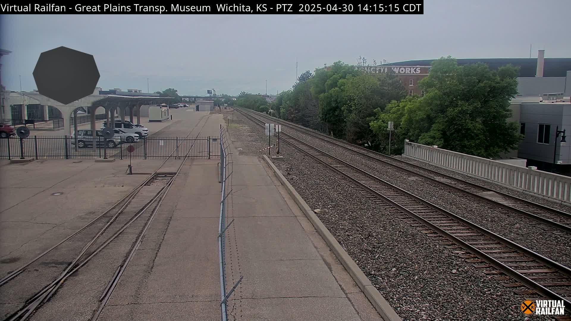 A mostly overcast day reveals a train yard with several parked cars near a building and parallel train tracks extending into the distance.