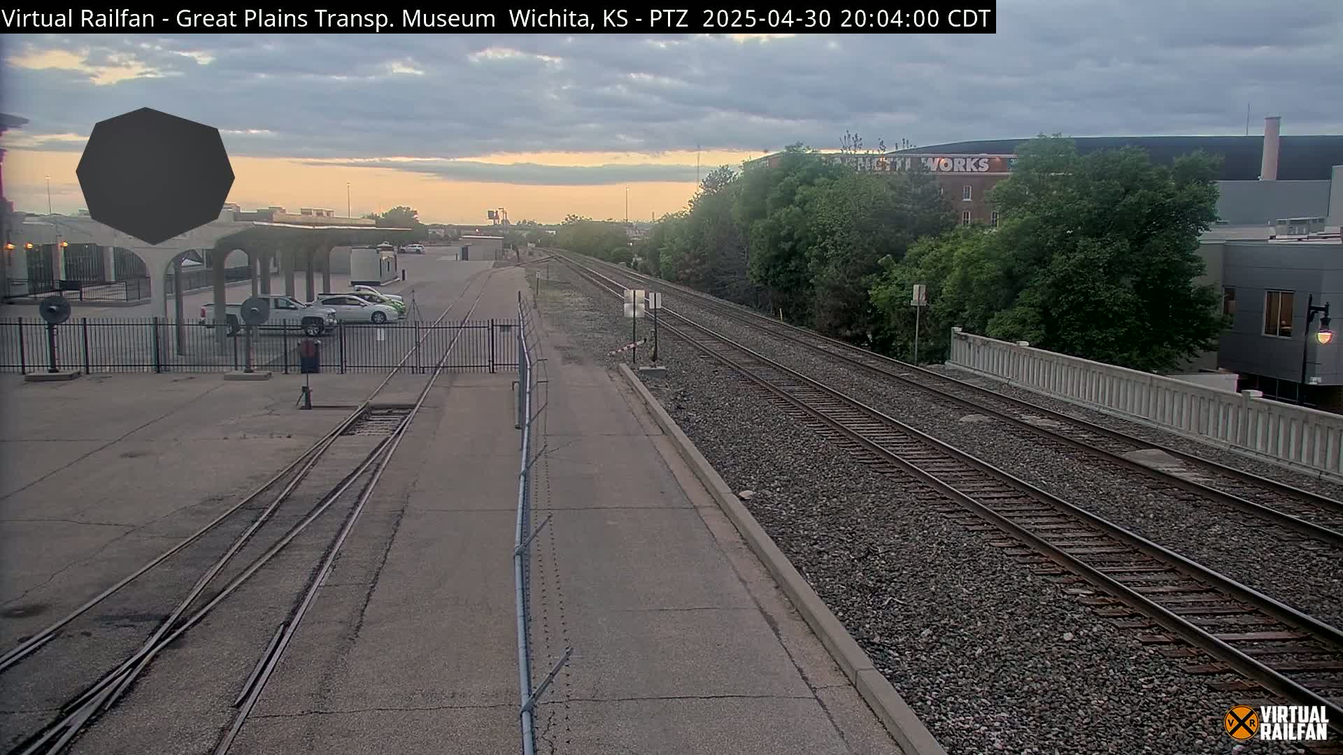 A dusk view of a railway switching yard with several parked cars, a fence, and trees beyond the tracks.