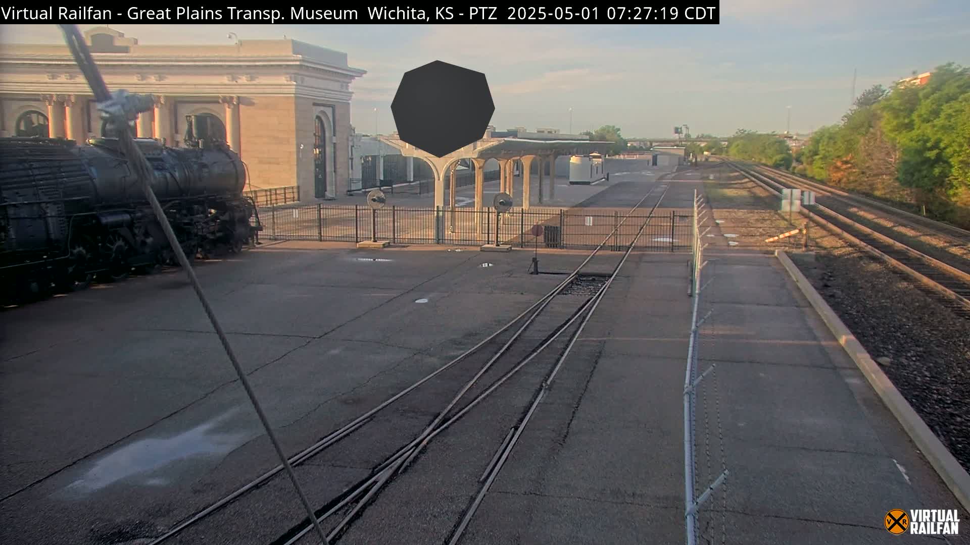 A large black steam locomotive sits near a museum building and railway tracks on a sunny morning.