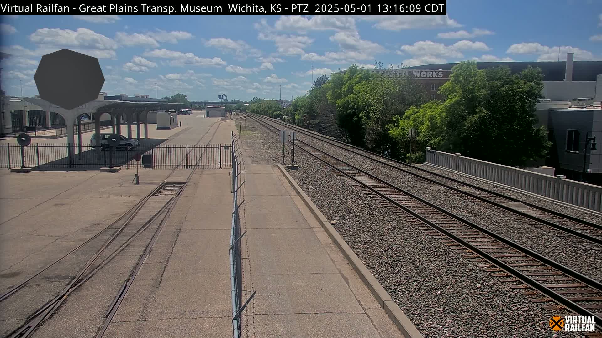 A sunny day reveals railroad tracks extending into the distance, passing alongside a fenced-off area with a covered platform, a parked car, and some buildings.