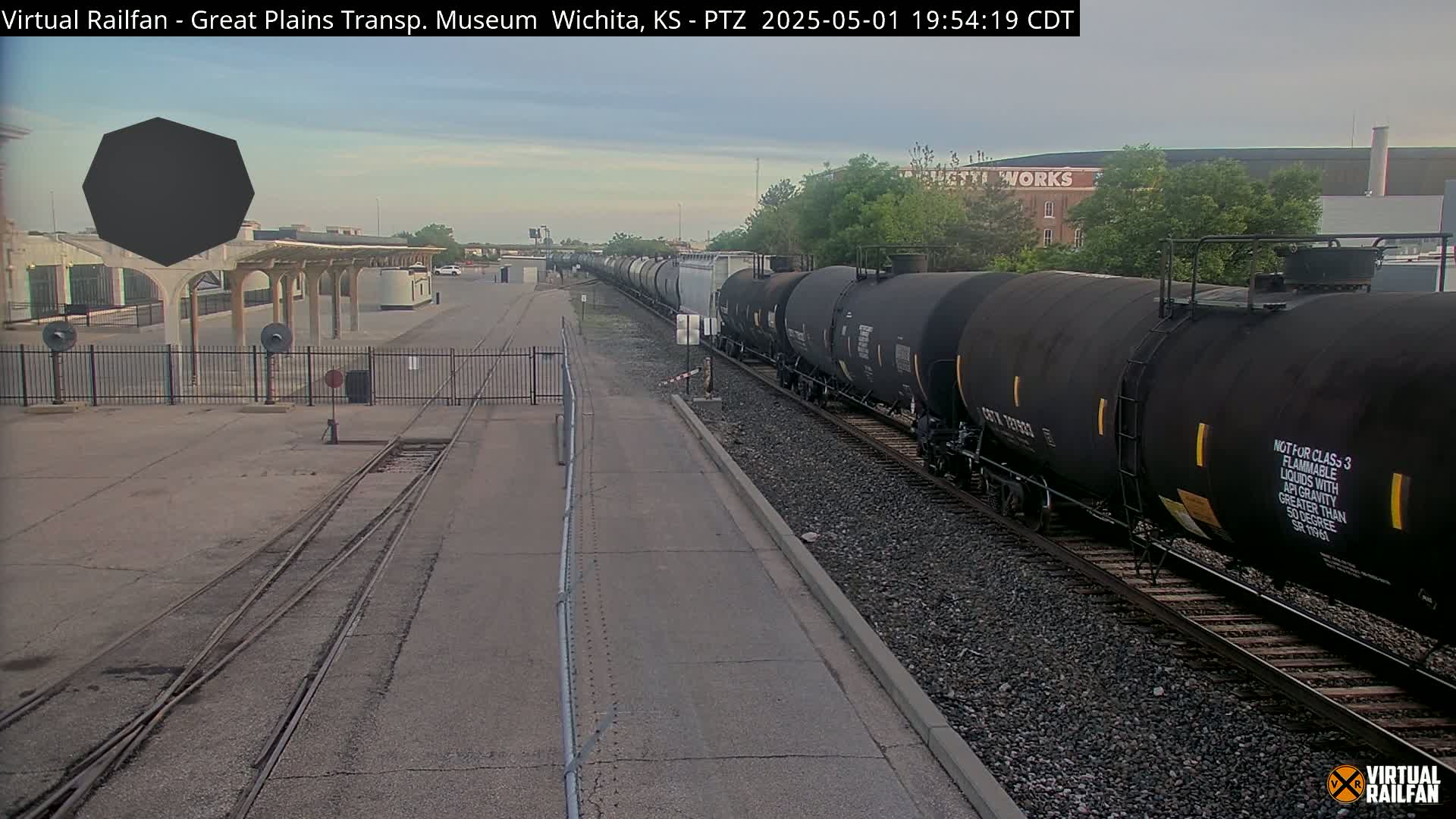 A long freight train of tanker cars passes by a rail yard under a partly cloudy sky.