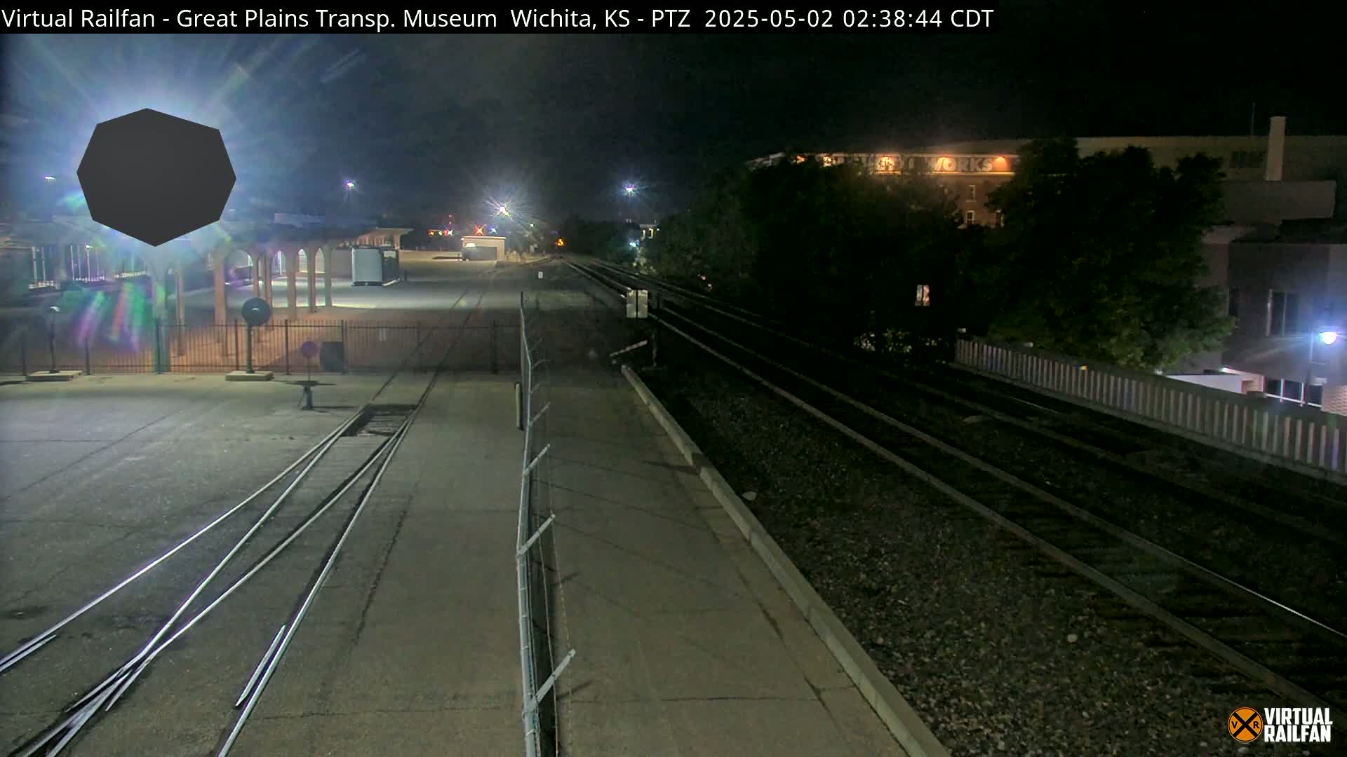 At night, a train track with a building and a fence in view, is illuminated under artificial light.