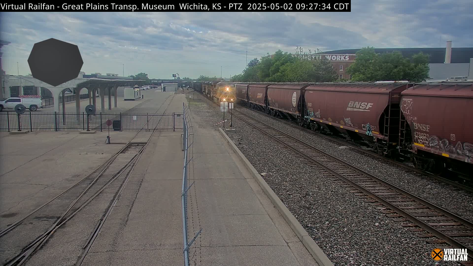 A long freight train passes by a railway station under a cloudy sky.