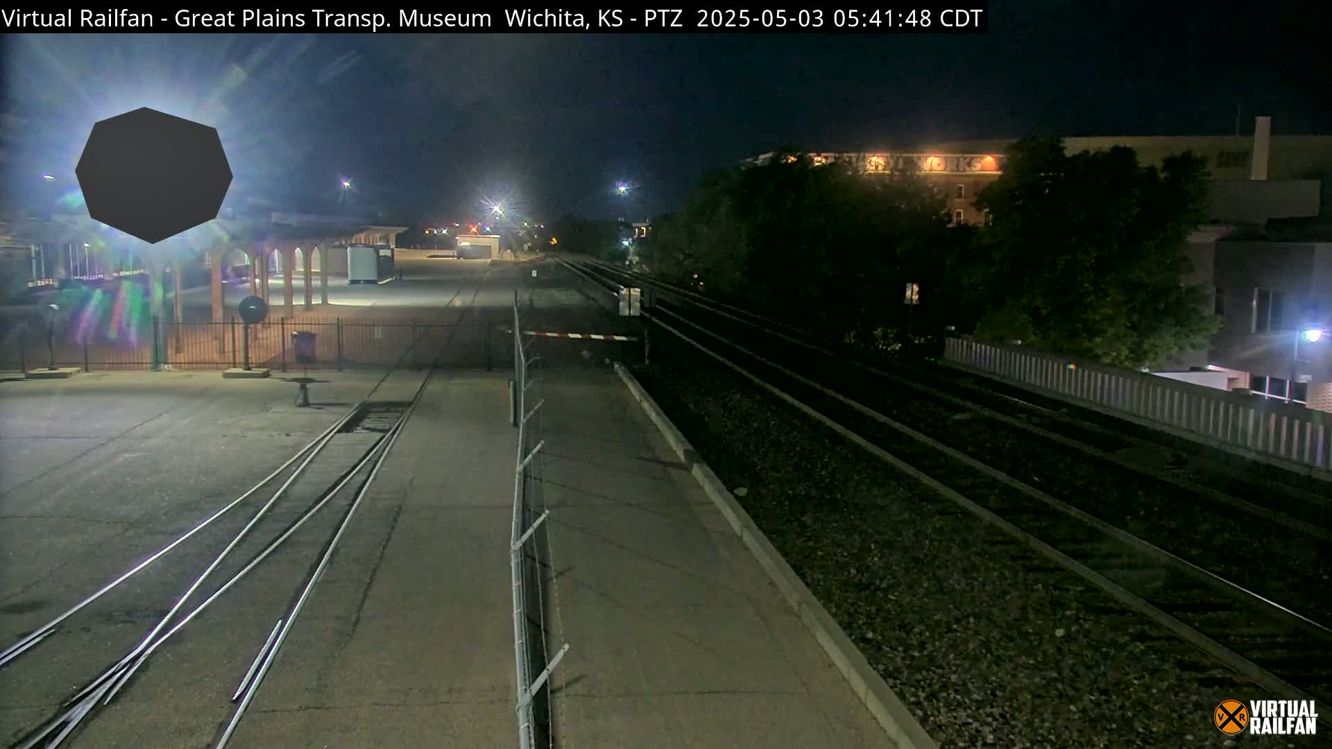 A nighttime view of train tracks extending into the distance, passing a fenced-in area with a building under a dark sky.