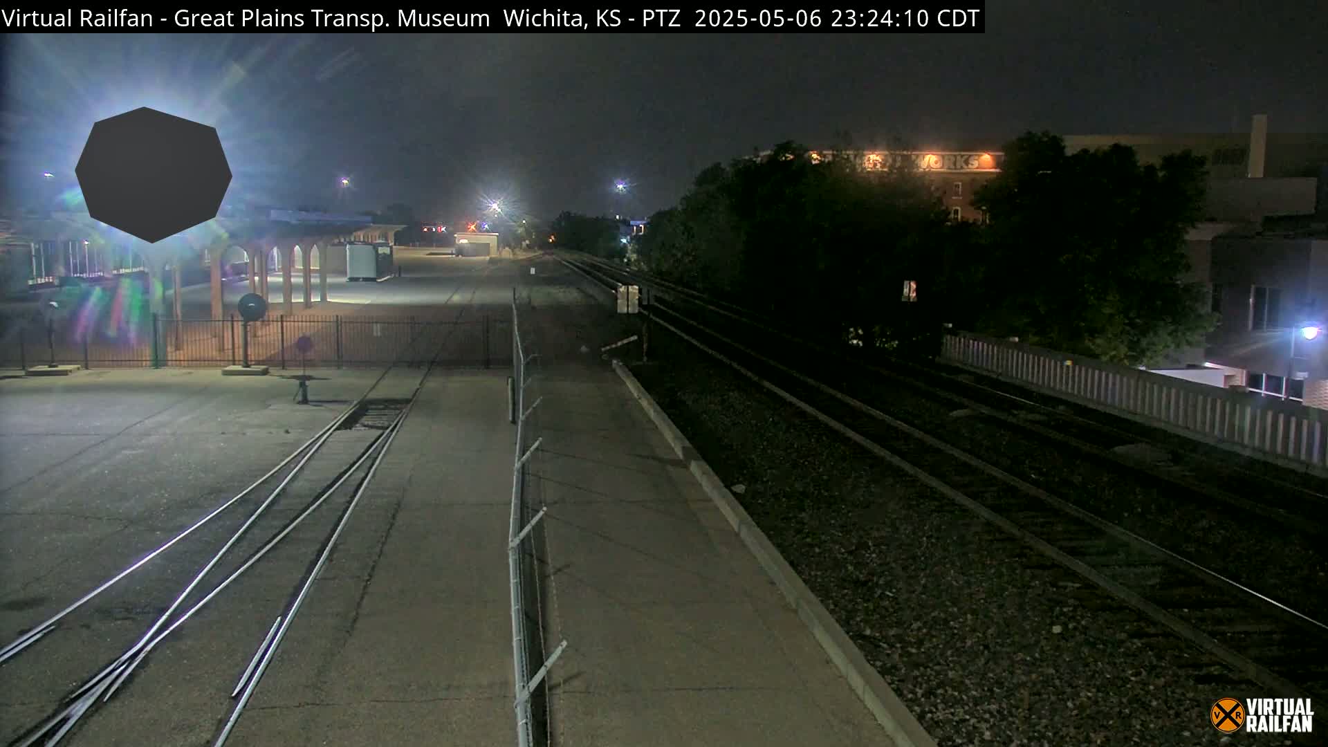 At night, railroad tracks extend from a dimly lit station area past a fence toward buildings and trees.