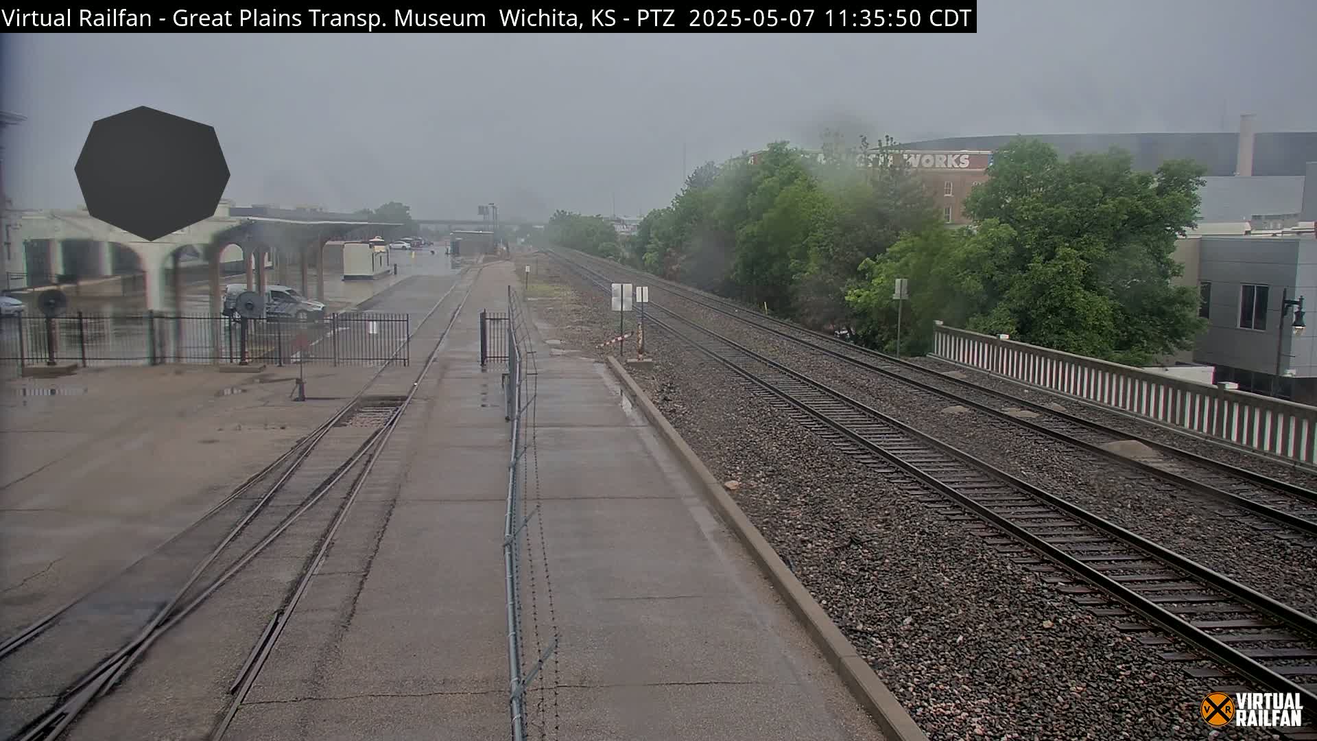 A wet train platform and railway tracks extend into the distance under an overcast sky.