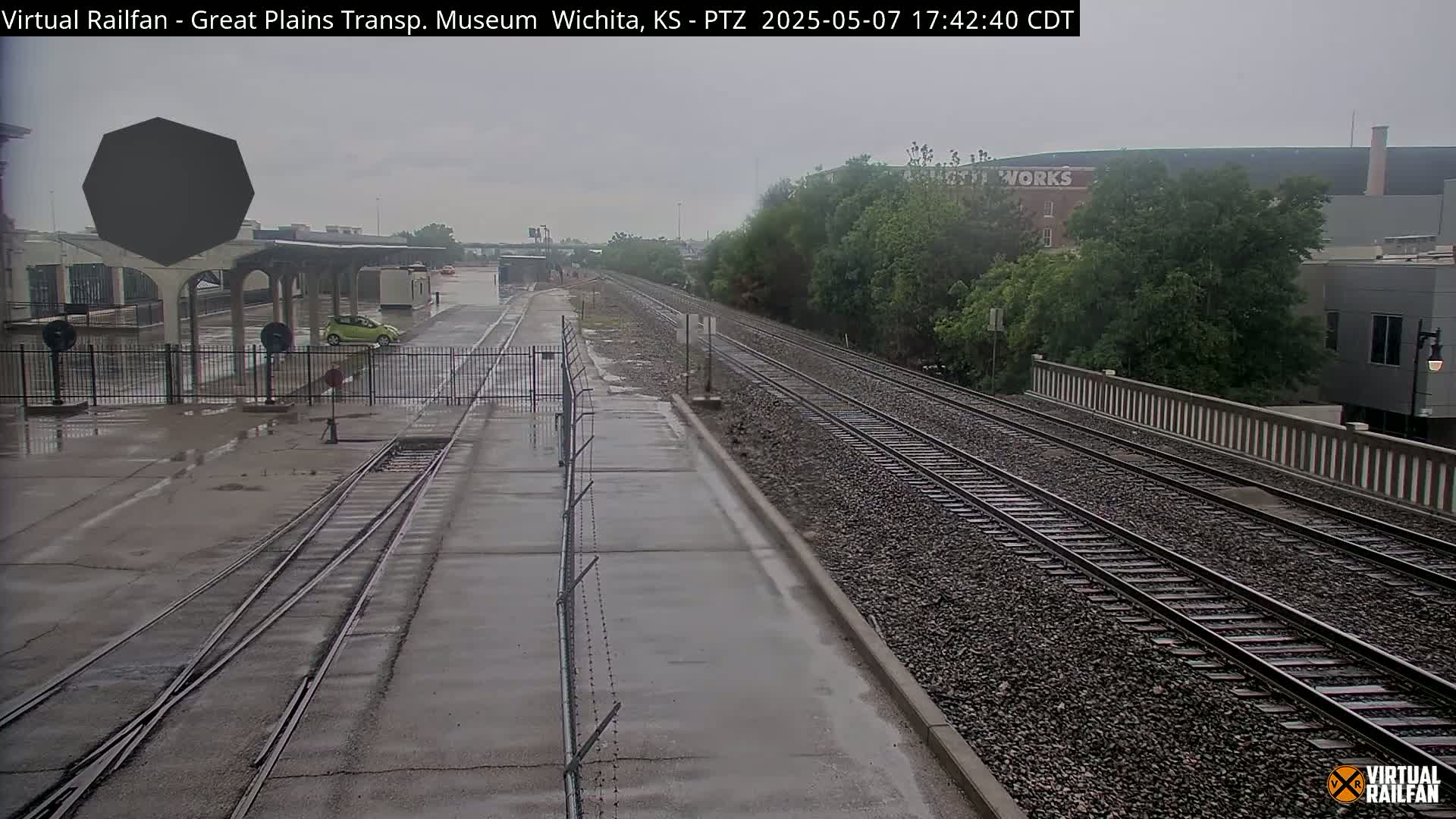 A wet, overcast day at a train station with a single car parked near a platform, and railroad tracks extending into the distance.