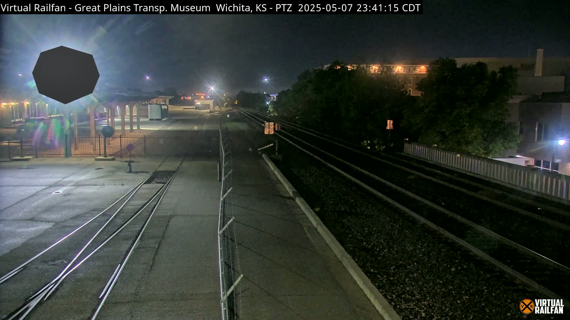 At night, a nighttime view shows train tracks extending into the distance past a chain-link fence, with a dark building and trees visible in the background.