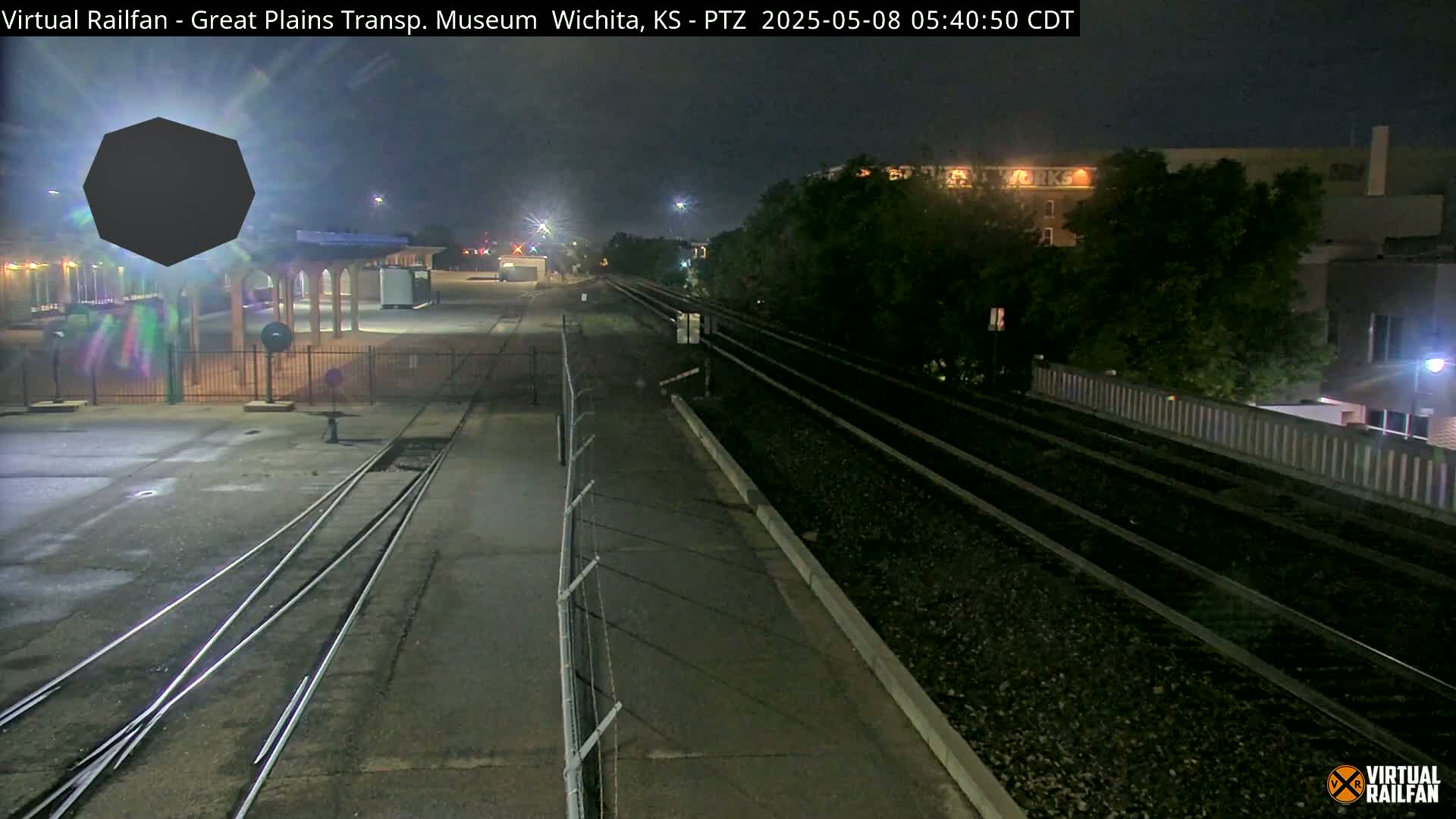 A nighttime view of train tracks extending into the distance, passing near a building and a fenced-in area with some structures, under a dark sky.