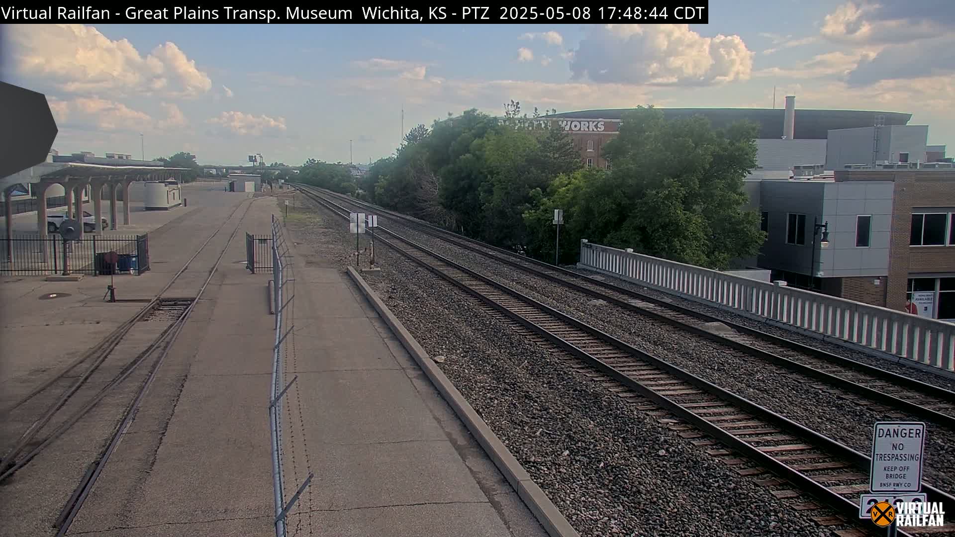 A partly cloudy sky overlooks a train station with railway tracks extending into the distance, passing under a bridge next to buildings and lush greenery.