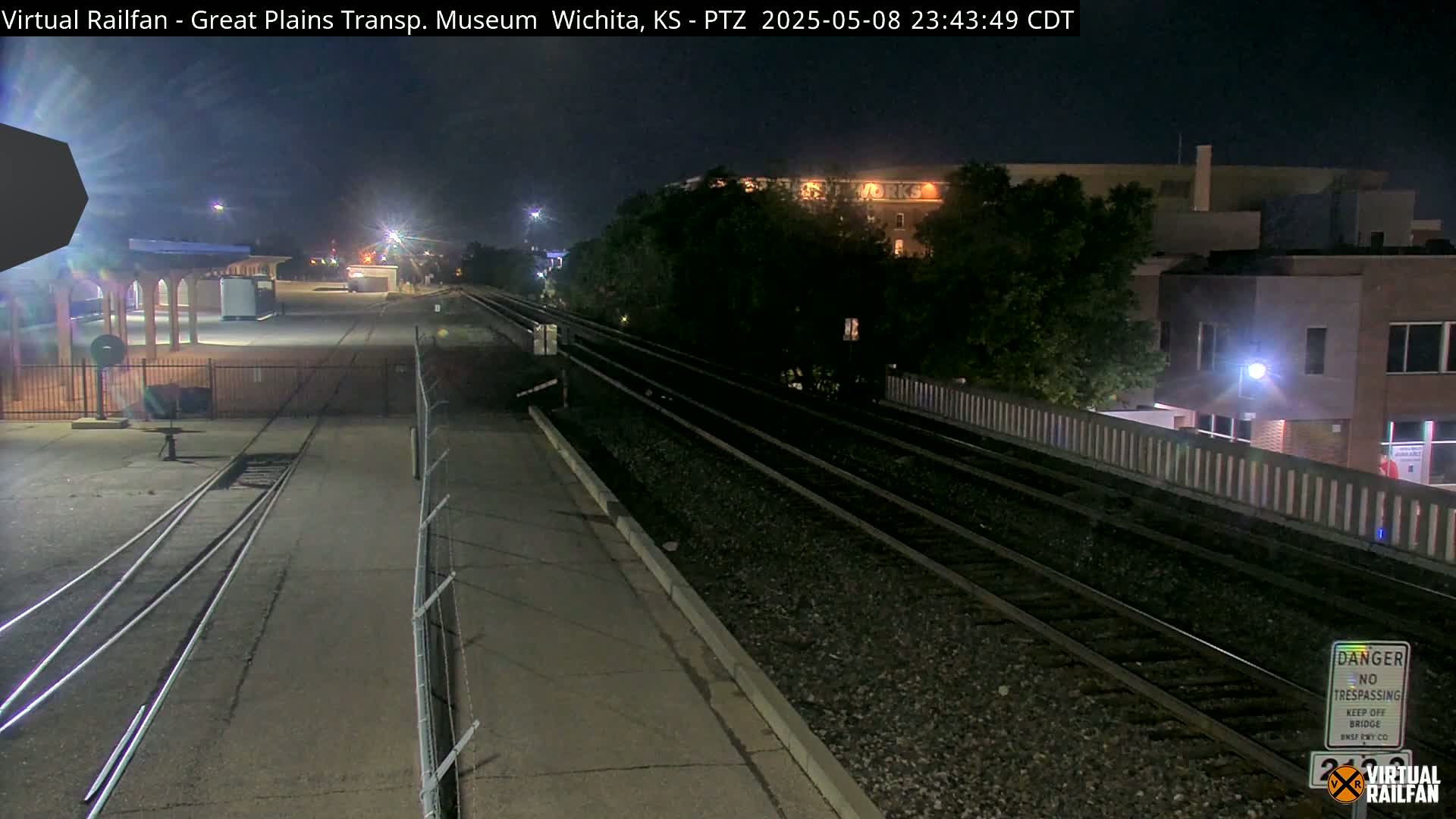 At night, train tracks stretch toward a building and trees under a clear sky.
