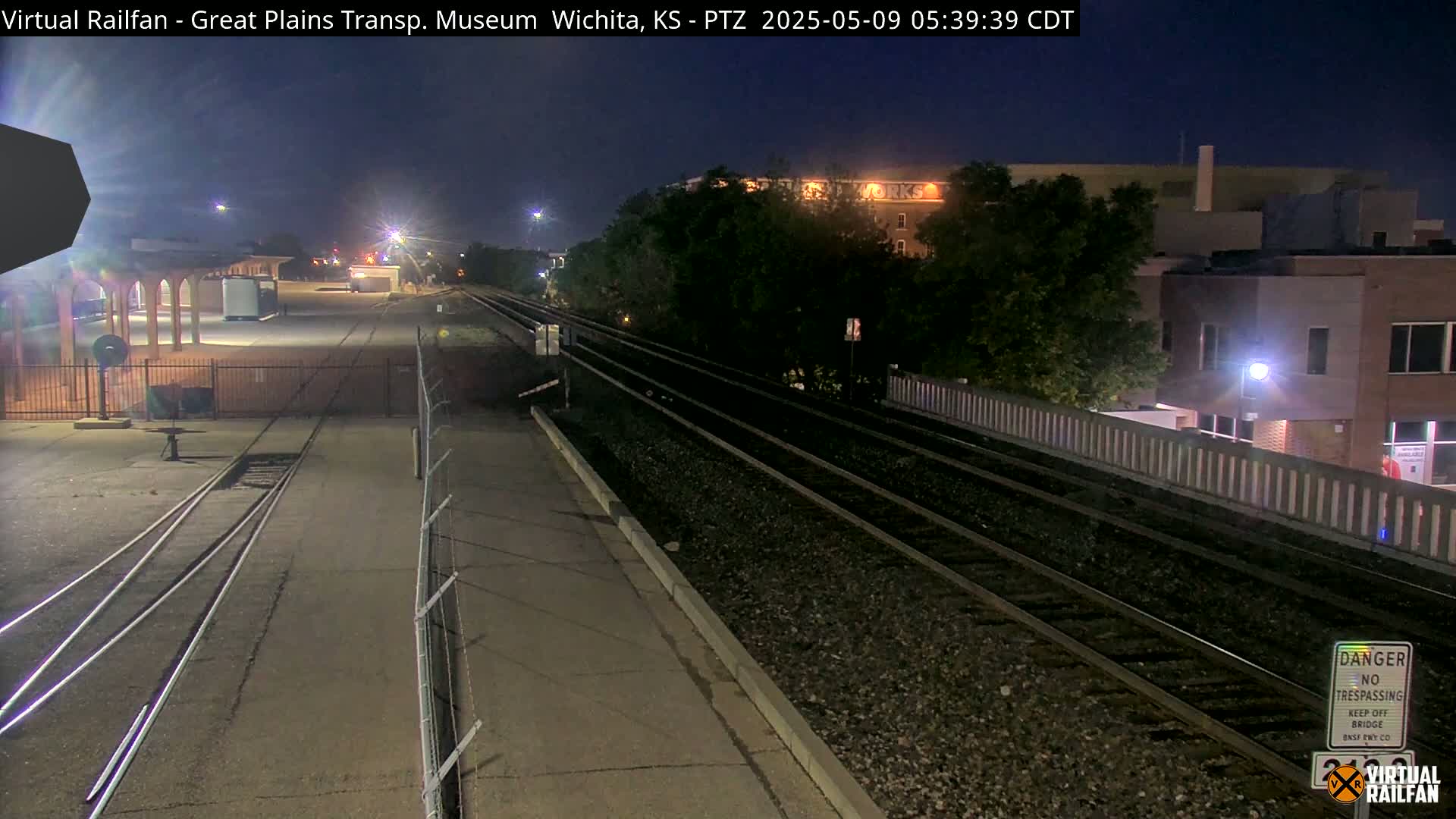 A nighttime view of train tracks beside a platform and fenced-off area, with buildings visible in the background under a dark sky.