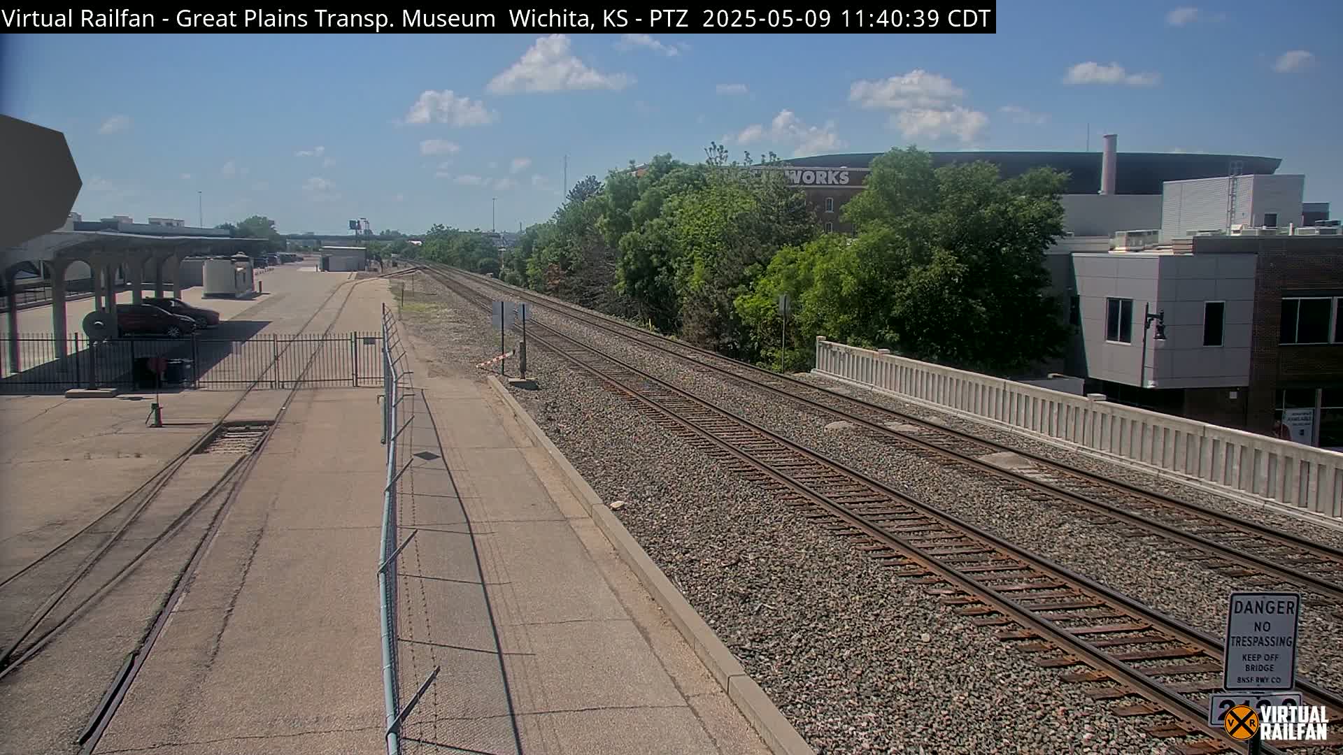 A sunny day reveals a view of railway tracks extending into the distance, passing alongside a fenced-off area with parked cars and a building in the background.