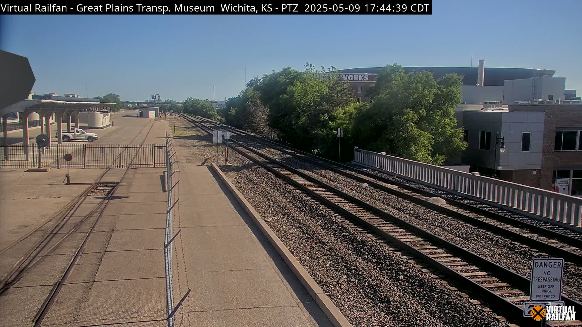 A sunny outdoor view of railroad tracks extending into the distance, passing by a fenced-in area and some buildings.