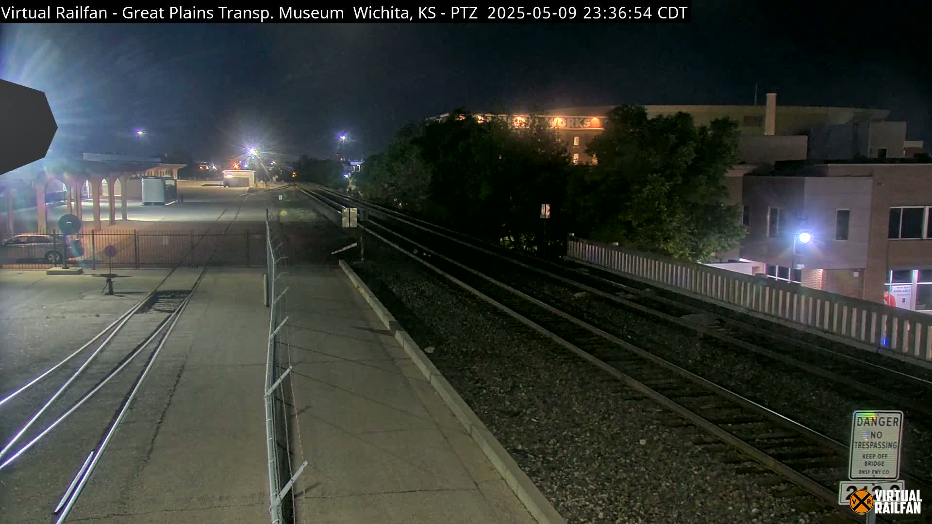 At night, train tracks extend into the distance past a fenced-in area next to a building, under a dark sky.