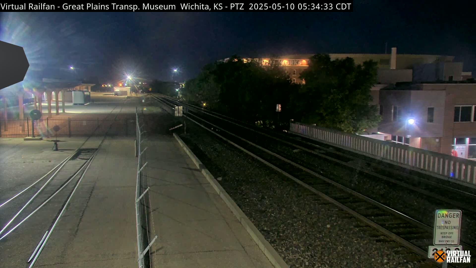 A nighttime view of train tracks extending into the distance past a fenced-in area and buildings, under a dark sky.