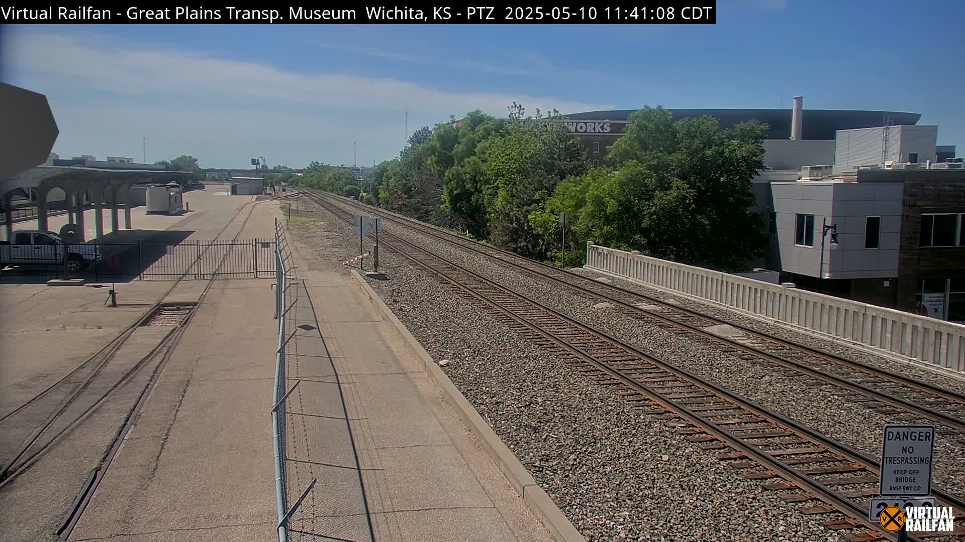 A sunny day shows a view of train tracks extending into the distance, beside a paved road and bordered by a chain-link fence, with buildings visible in the background.