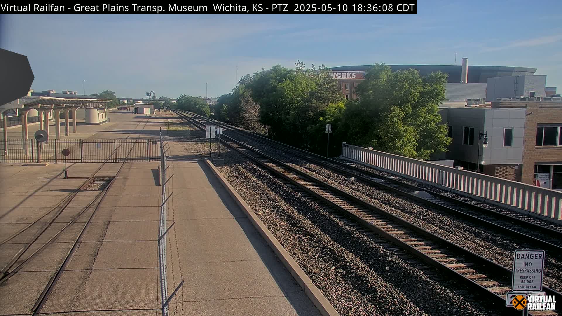 A sunny view of railroad tracks extending into the distance, passing alongside a low fence and a building, with trees and other structures visible in the background.